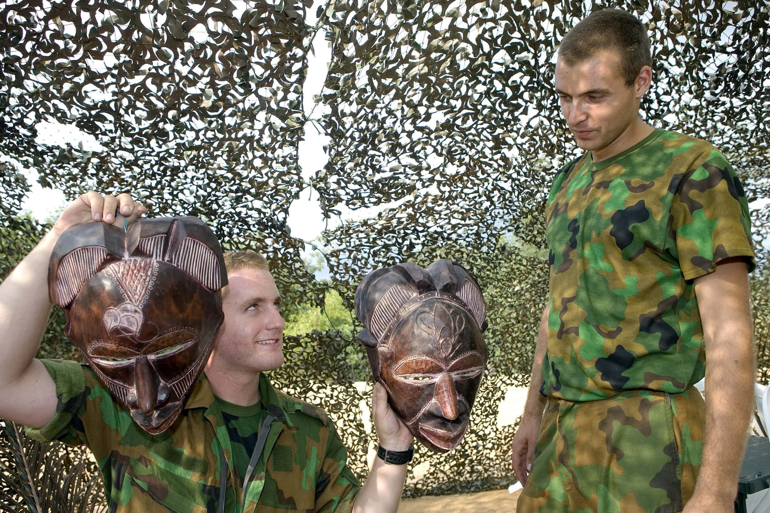 Kleurenfoto, twee mannen in camouflagekleding onder camouflagenetten, de man links houdt twee houten maskers omhoog.