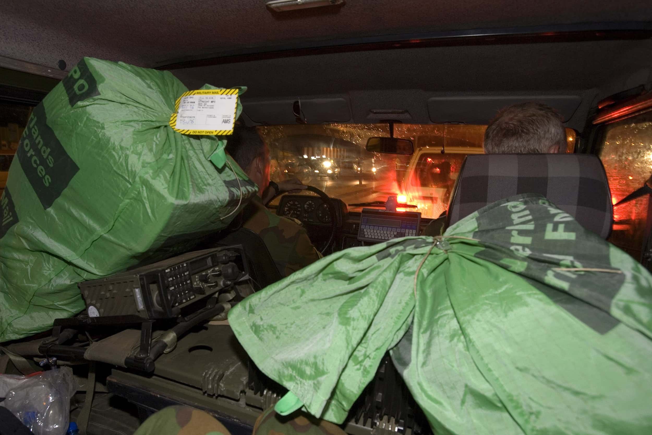 Kleurenfoto, twee mannen zitten voorin in een auto, op de voorgrond twee grote groene zakken.