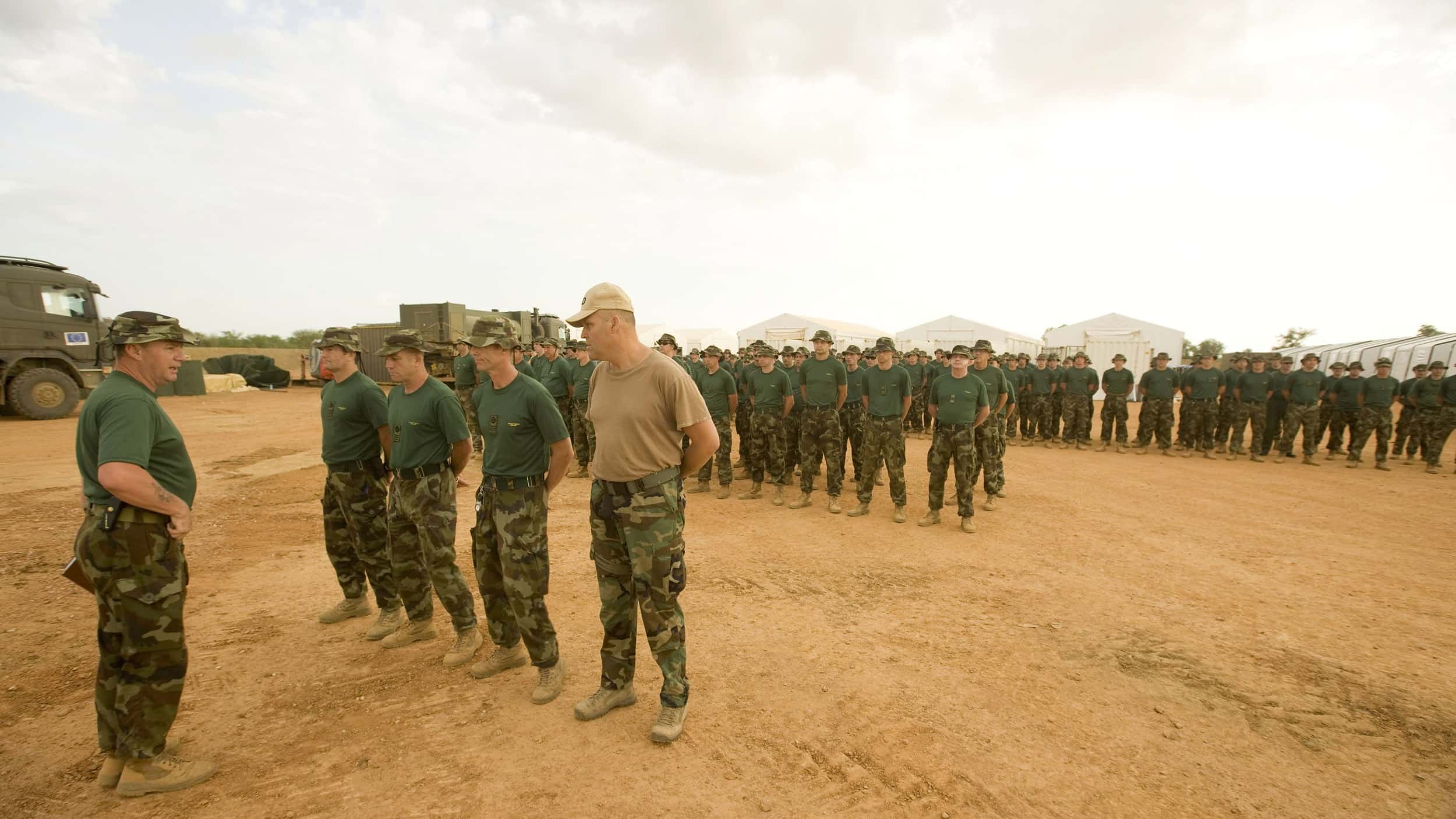 Kleurenfoto. Op een zandvlakte in de buitenlucht staan tientallen militairen in rijen opgesteld. Ze dragen broeken in camouflagekleuren, een groenblauw shirt en een zonnehoed. In de achtergrond staan vrachtwagens en zijn de bovenkanten van witte tenten te zien.