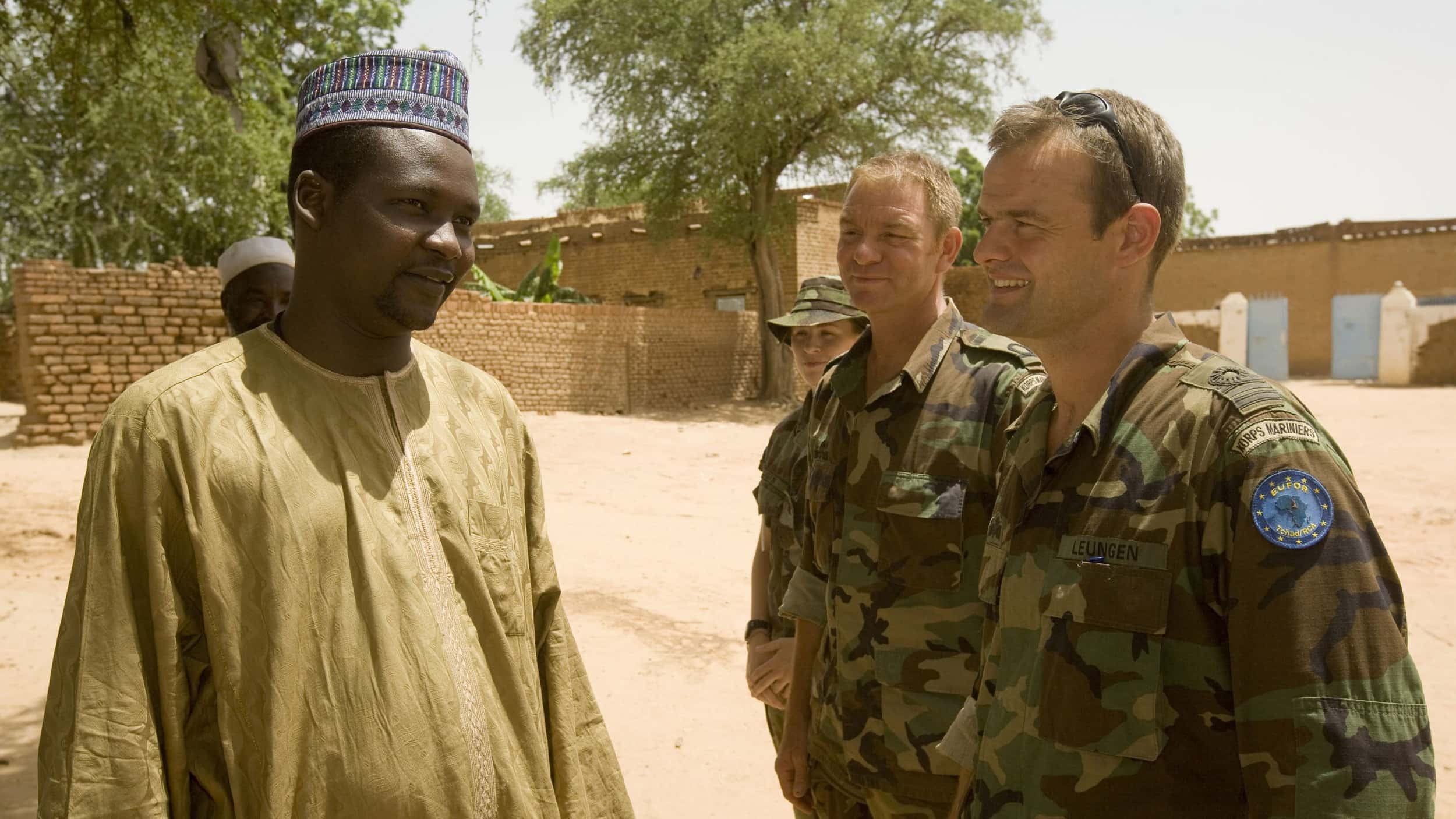 Kleurenfoto. Rechts staan drie militairen, gekleed in uniformen met camouflagekleuren. De meest rechter van hen heeft een zonnebril in het haar. Ze zijn in gesprek met een man links, hij is gekleed in een donkergeel gewaad, hij draagt een groen-paars mutje. In de achtergrond bakstenen muren en bomen.