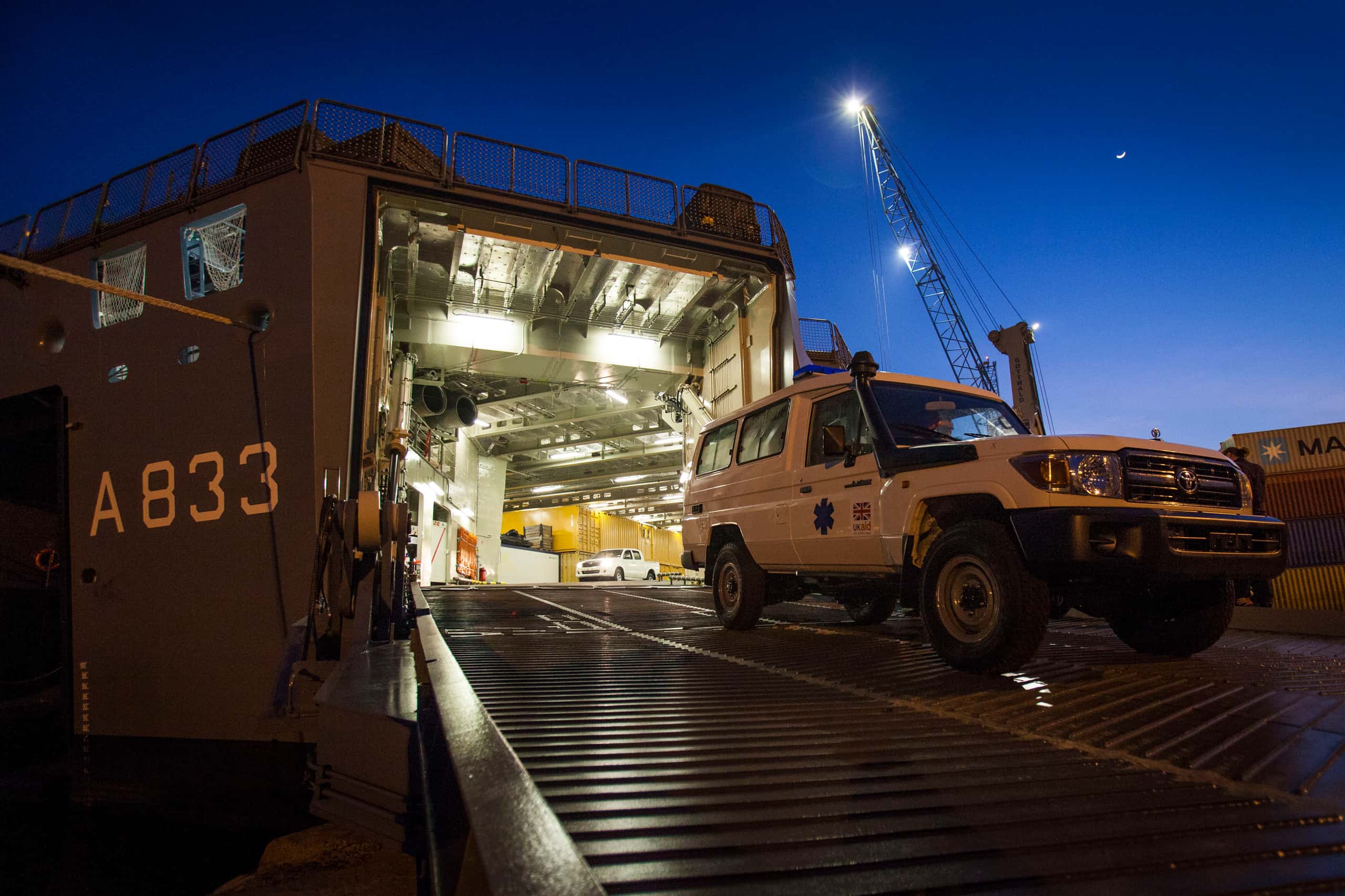 Kleurenfoto. Te zien is de voorkant van een schip die open staat. Links op de boeg staat A833 geschreven. Een laadklep staat omlaag, binnen in het laadruim brandt ligt. Op de laadklep rijdt een witte auto, in het ruim is nog een witte auto te zien. Buiten is het donker, aan een kraan branden twee lampen, de maan schijnt.
