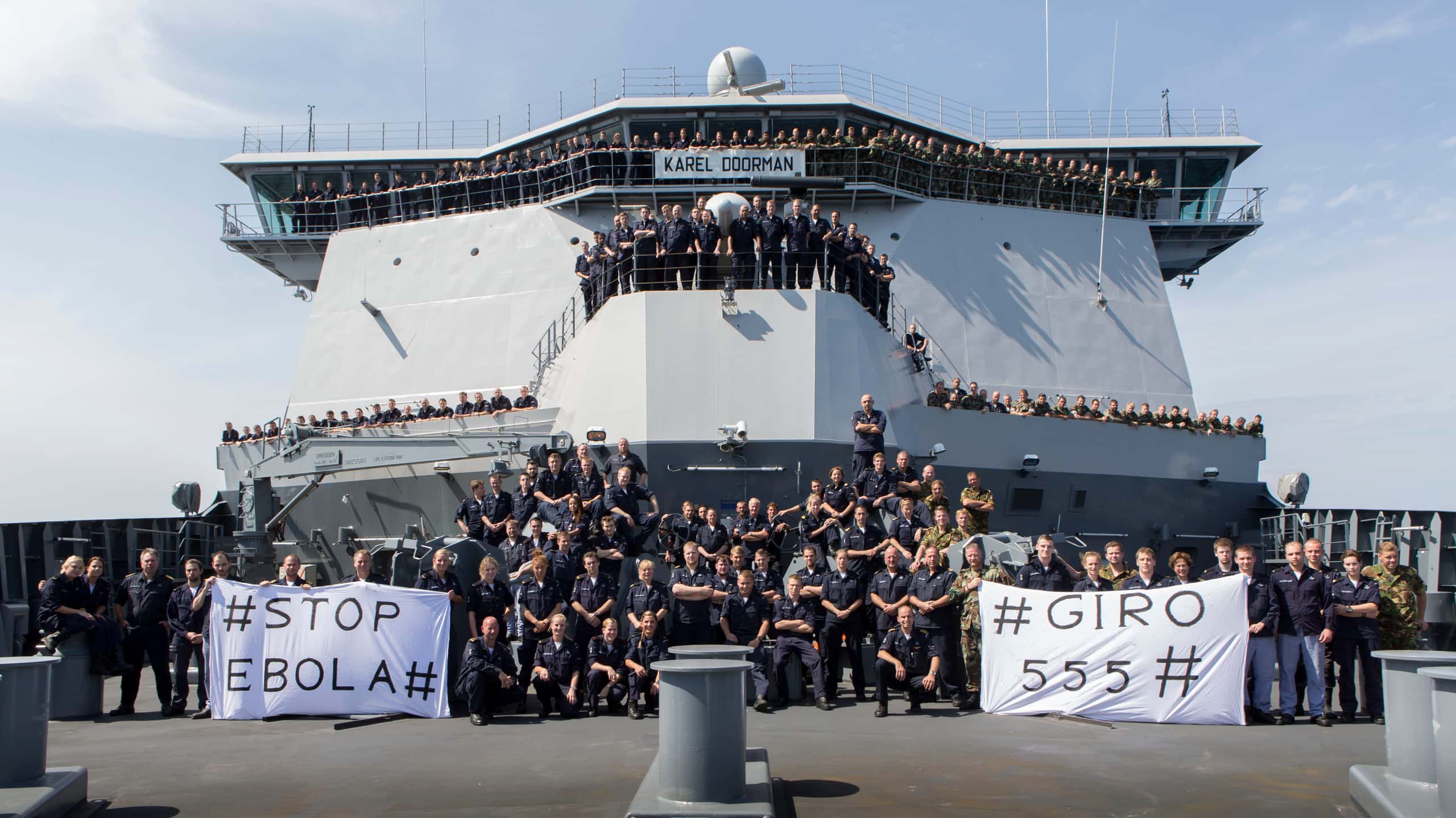 Kleurenfoto. Groepsfoto van tientallen militairen, die voor, op en rond de brug van een marineschip poseren. Het merendeel draagt donkerblauwe uniforms, anderen een uniform in camouflagekleuren. De militairen op het dek houden twee spandoeken vast, eentje links en eentje rechts. Links staat #stop ebola# en rechts #giro 555#.