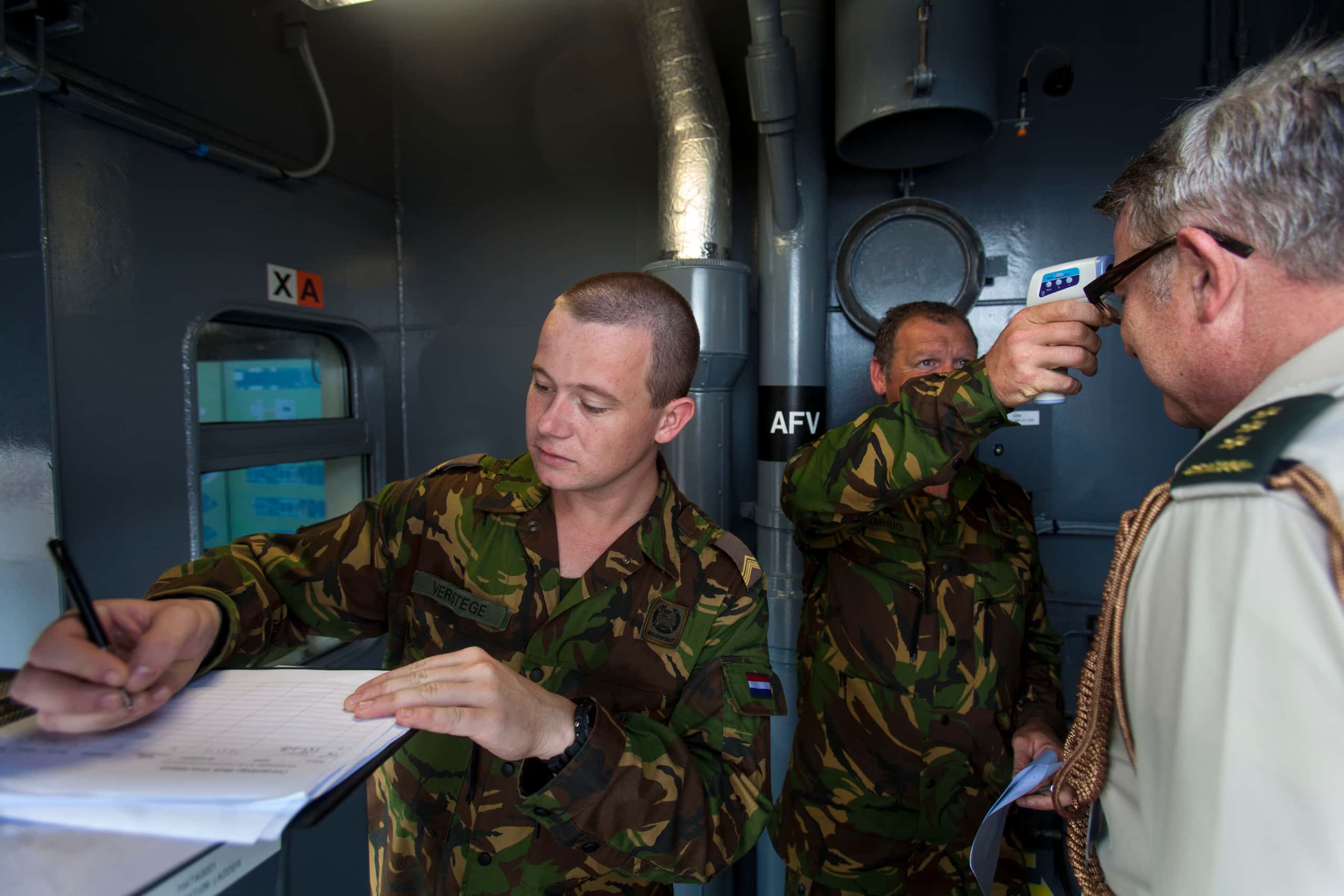 Kleurenfoto. Drie militairen staan in een kleine ruimte. De middelste van hen houdt een thermometer in de vorm van een pistool tegen het voorhoofd van de militair rechts. De militair links staat met zijn rug naar de andere twee en schrijft iets op. De middelste en linker militair dragen een uniform in camouflagekleuren.