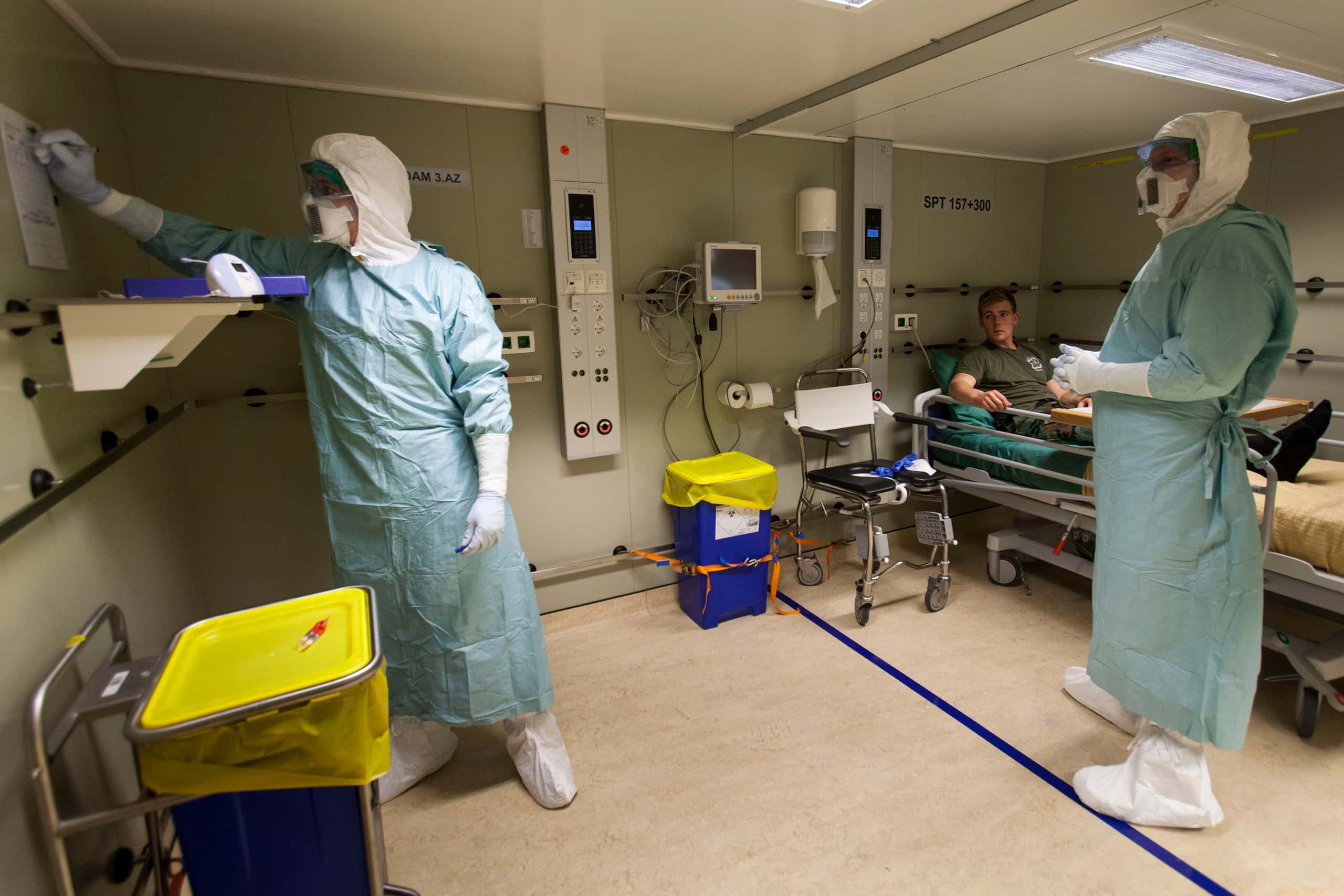 Kleurenfoto. Te zien is een ruimte die als ziekenboeg is ingericht. Rechts tegen de muur staat en ziekenhuis bed waar een jongeman in ligt, aan de muur hangen stopcontacten, knoppen en schermpjes. In de ruimte staan nog twee andere personen, die van top tot teen in blauw en wit plastic gekleed zijn, inclusief mondkapjes en veiligheidsbrillen. De linker van hen schrijft iets op een papier dat aan de muur hangt.
