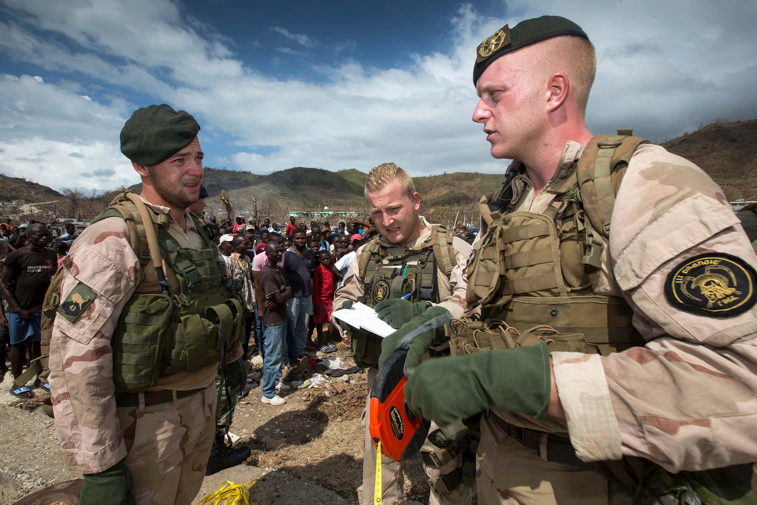 Kleurenfoto. Drie militairen staan, in vol ornaat gekleed, met elkaar te praten. Ze kijken vermoeid. De rechter draagt een baret en rolt een meetlint op, de middelste schrijft iets op, de linker draagt ook een baret. Achter hen is een grote groep burgers te zien. In de achtergrond een kaal heuvellandschap.