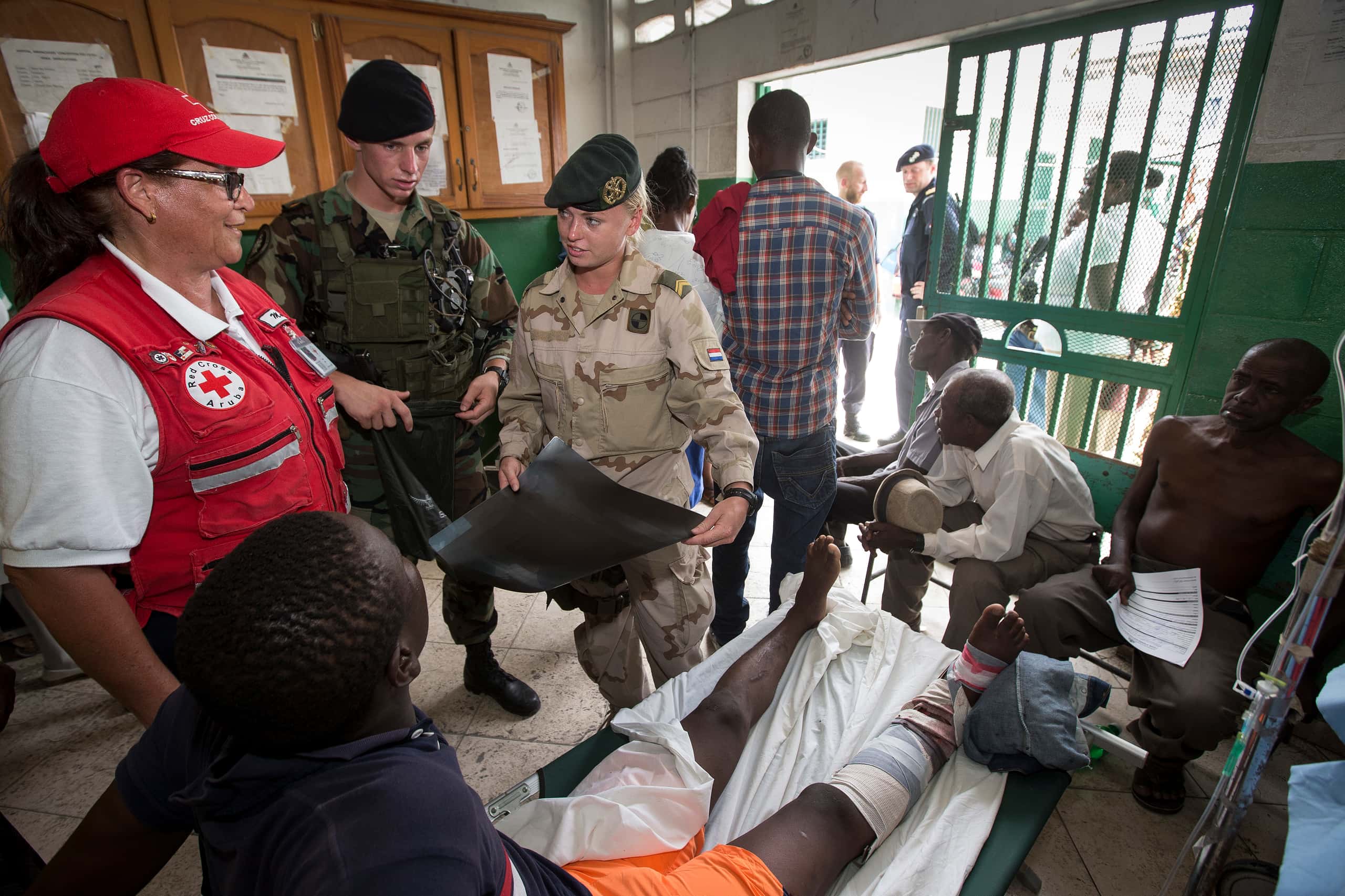 Kleurenfoto. Twee militairen zijn in een ziekenhuiskamer in gesprek met een Rode Kruismedewerker. Beide militairen dragen camouflagekleding en een baret, één van hen heeft een röntgenfoto vast. De Rode Kruismedewerker draagt een rode vest en rode pet. Op de voorgrond ligt een man met verband om zijn rechterbeen op een veldbed. In de hoek tegen de muur, in de deuropening en buiten de kamer zijn meer personen te zien.