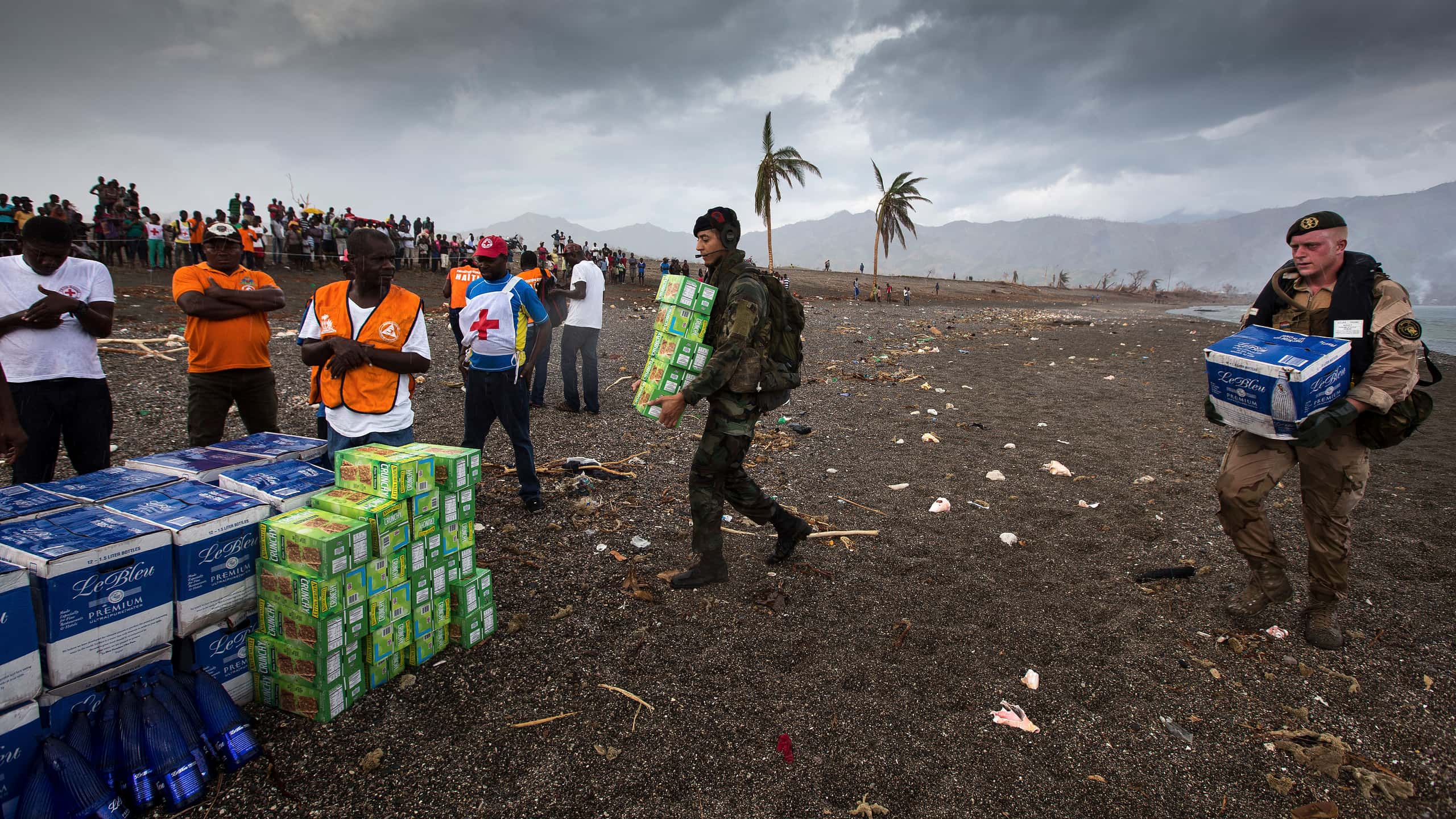 Kleurenfoto. Rechts twee militairen die op een strand allebei dozen dragen naar een verzameling van dezelfde dozen links op de foto. Naast de verzameling staan enkele mannen, zij dragen T-shirts of hesjes met een rood kruis erop. Links in de achtergrond staat een grote groep mensen achter een lint toe te kijken, verderop een enkele palmboom, helemaal rechts is de branding van de zee te zien. Verderop in de achtergrond bergen.