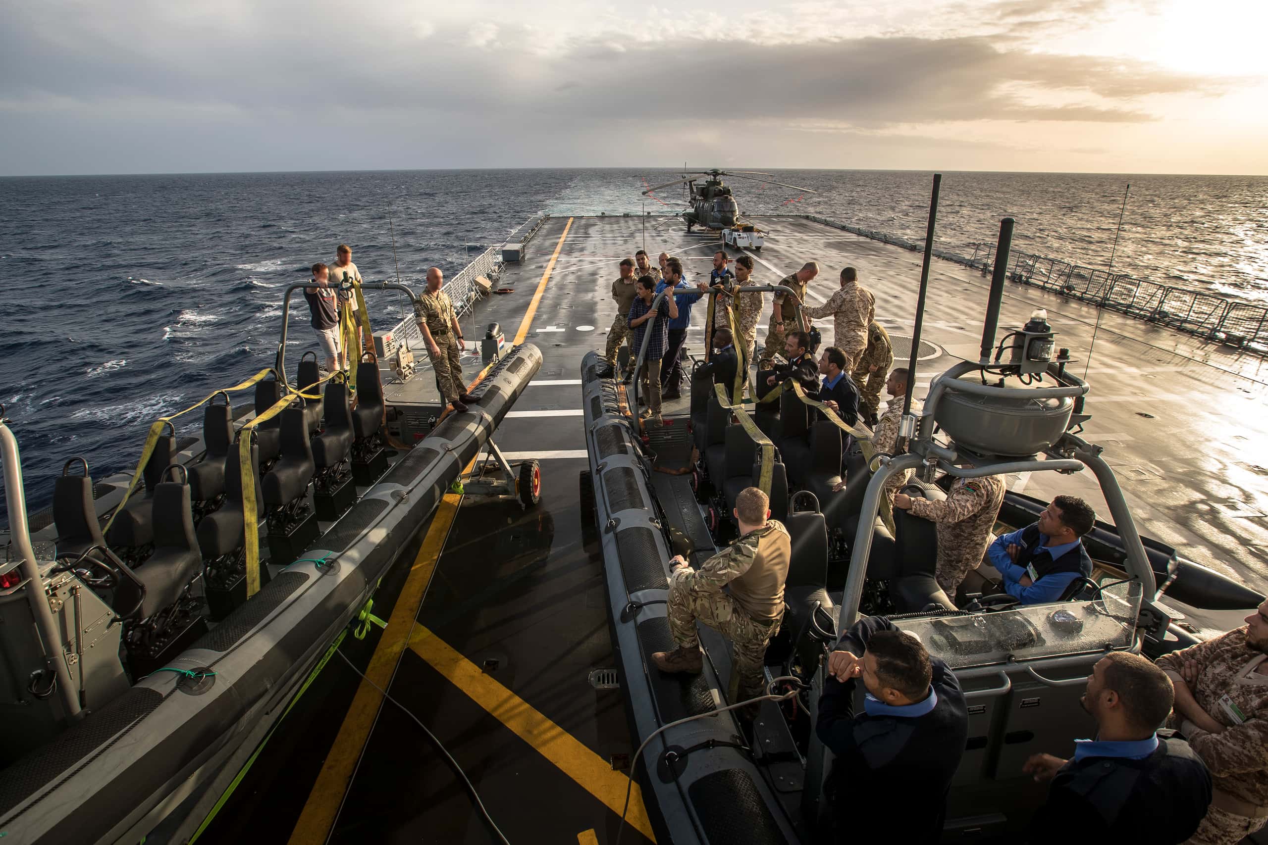 Kleurenfoto, op het dek van een schip. Er zijn twee rubberboten op een scheepsdek te zien, ze staan op wielen. Beide boten hebben twee rijen stoelen in het midden. Op de linker boot staan drie militairen, op de rechter boot staan er ongeveer twintig. De twintig op de rechter boot luisteren aandachtig naar de drie op de linker boot. Verderop op het dek een helikopter.