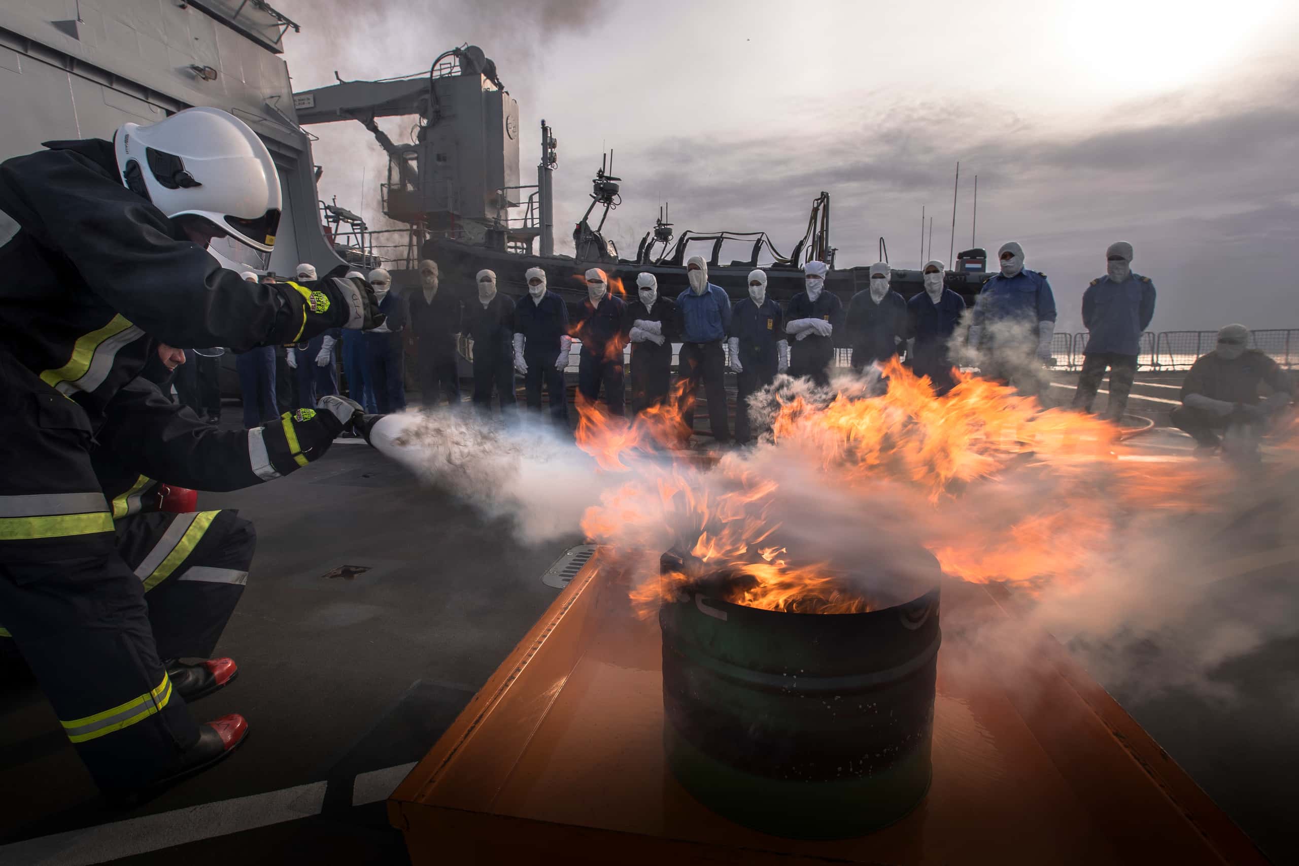 Kleurenfoto, op het dek van een schip. Links staan twee brandweermannen, in vol ornaat, die een vuur in een ijzeren vat blussen. Er zijn veel vlammen en rook. In de achtergrond staat een rij van zeventien personen toe te kijken, zij dragen witte bivakmutsen en lange witte handschoenen.