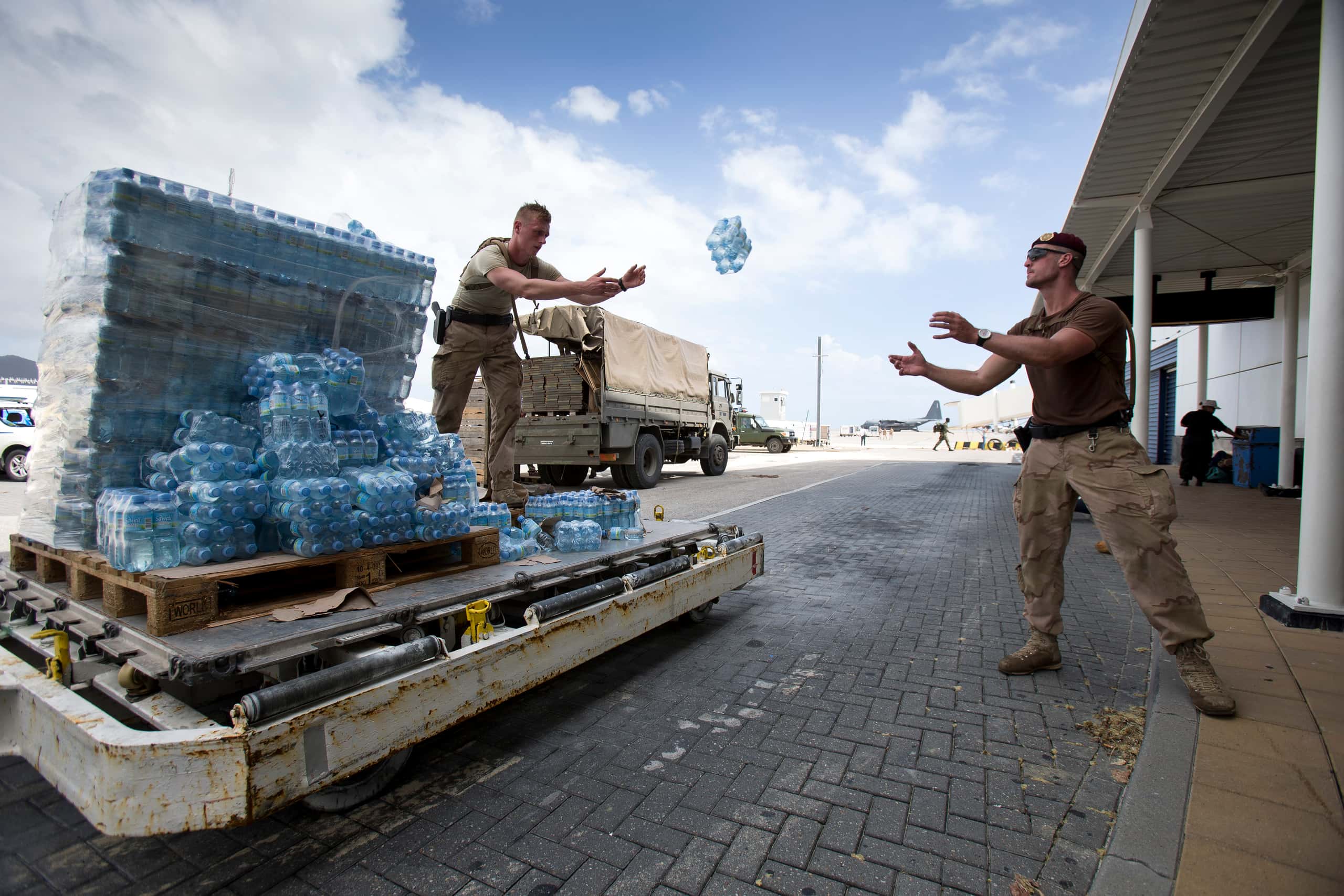Kleurenfoto. Twee militairen staan op een parkeerplaats. Helemaal rechts is een overkapping. De linker van de twee staat op een palet met tientallen pakken flessen water en gooit een pak naar de ander. In de achtergrond een legergroene vrachtwagen.
