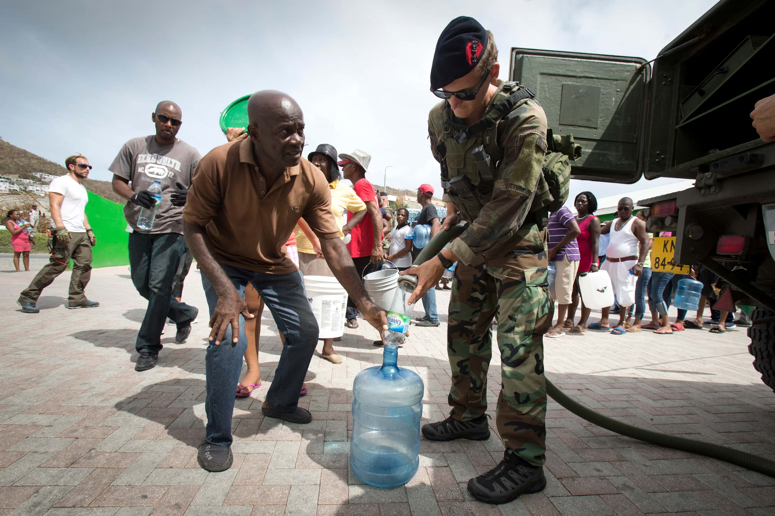 Kleurenfoto. Een militair houdt een waterslang vast en laat water in een grote plastic fles lopen. Naast hem staat een man in burgerkleding, die een klein flesje aan de tuit van de grote fles houdt, als een trechter. Achter de man begint een lange rij van mensen die flessen en emmers vast hebben. Helemaal rechts, naast de militair, is de achterkant van een militair voertuig half te zien.