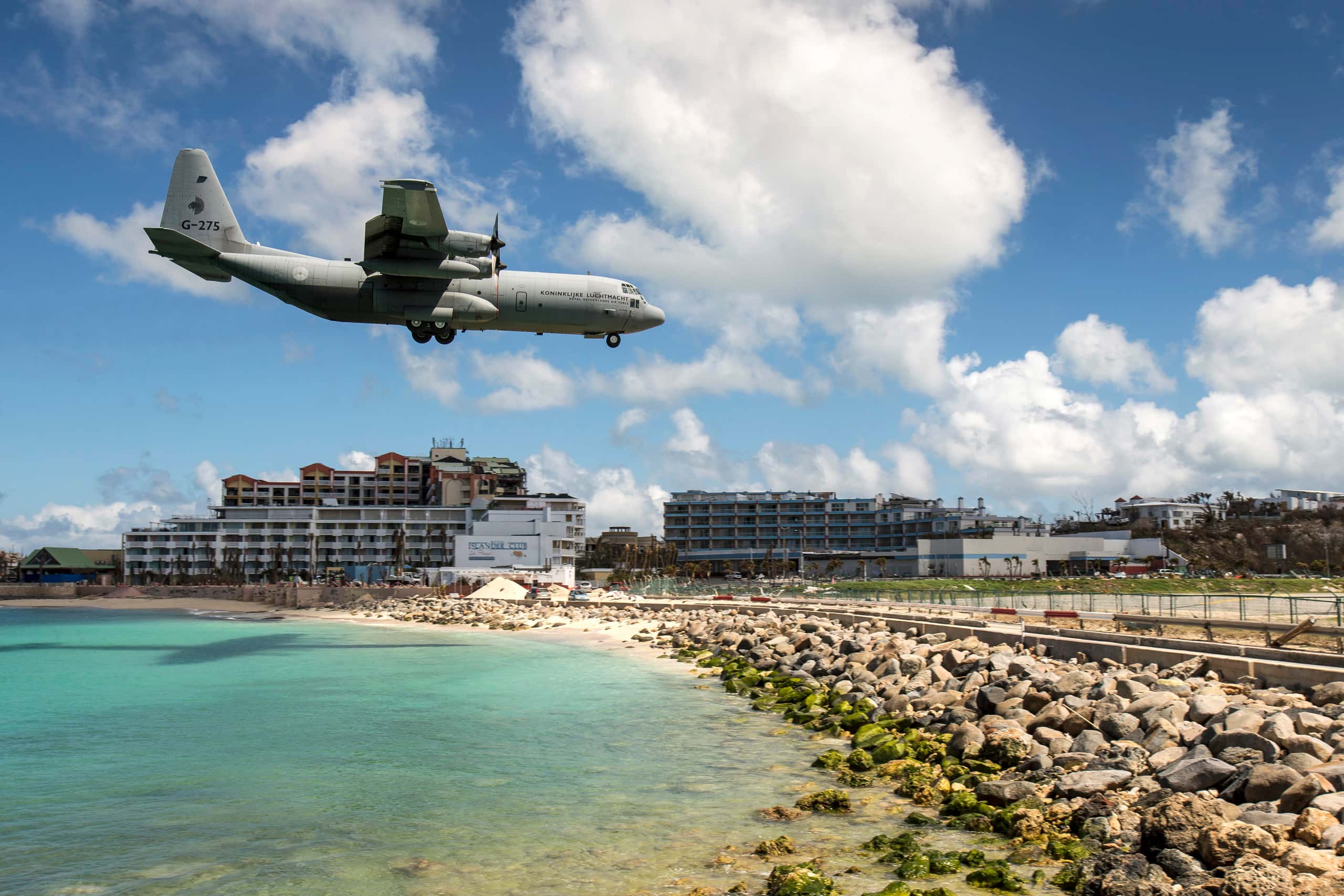 Kleurenfoto. Een groot, grijs vliegtuig hangt in de lucht. Links is een stuk zee te zien. Rechts begint het land, met rotsblokken en daarachter meteen het begin van de landingsbaan. In de achtergrond twee grote hotels.