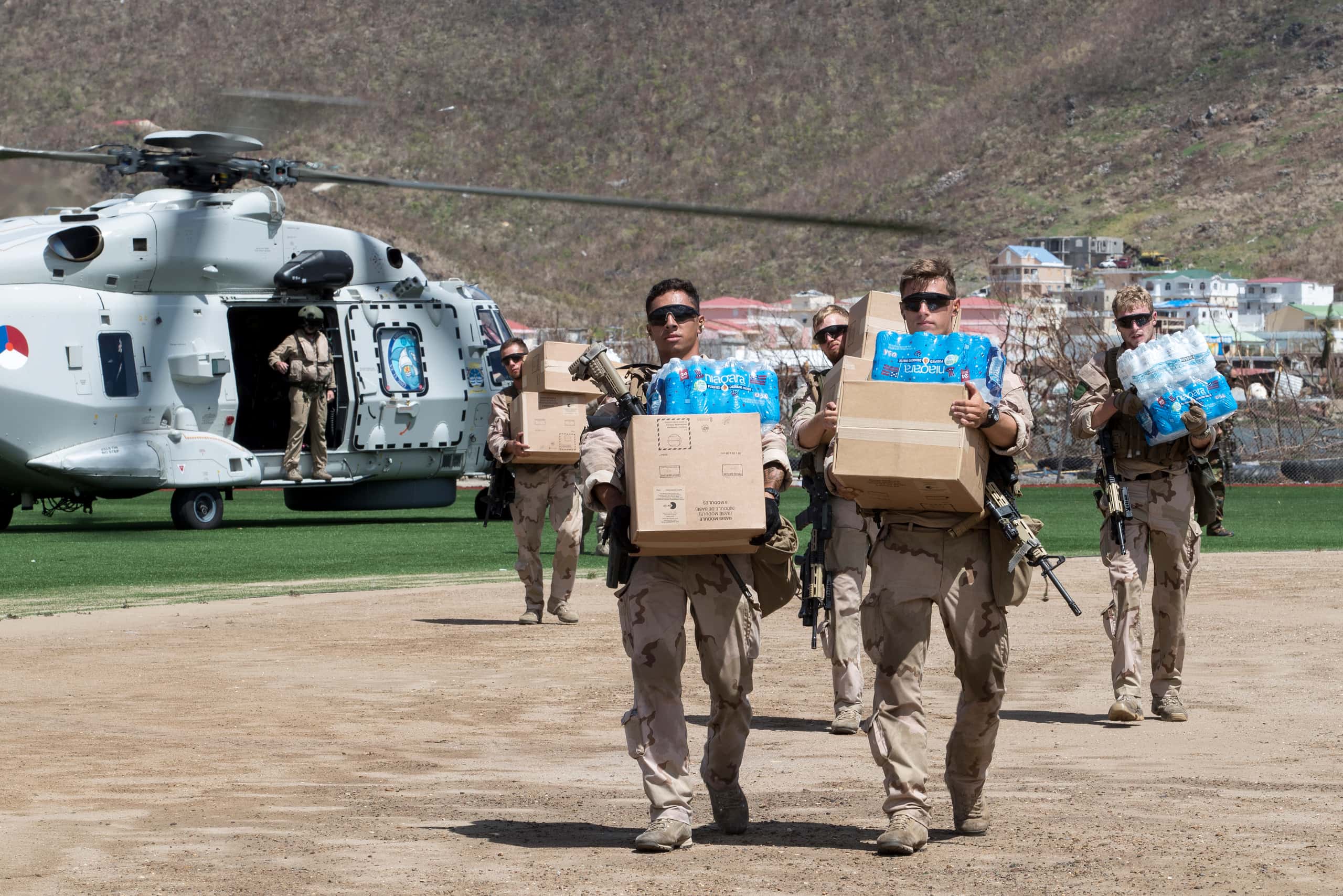 Kleurenfoto. Vijf militairen lopen weg van een helikopter, die nog met draaiende propellers links in de achtergrond staat. De militairen dragen beige uniformen en zonnebrillen. Elk van hen draagt bovendien één of twee kartonnen dozen en/of pakket drinkwater. Hun geweren steken onder de dozen uit.