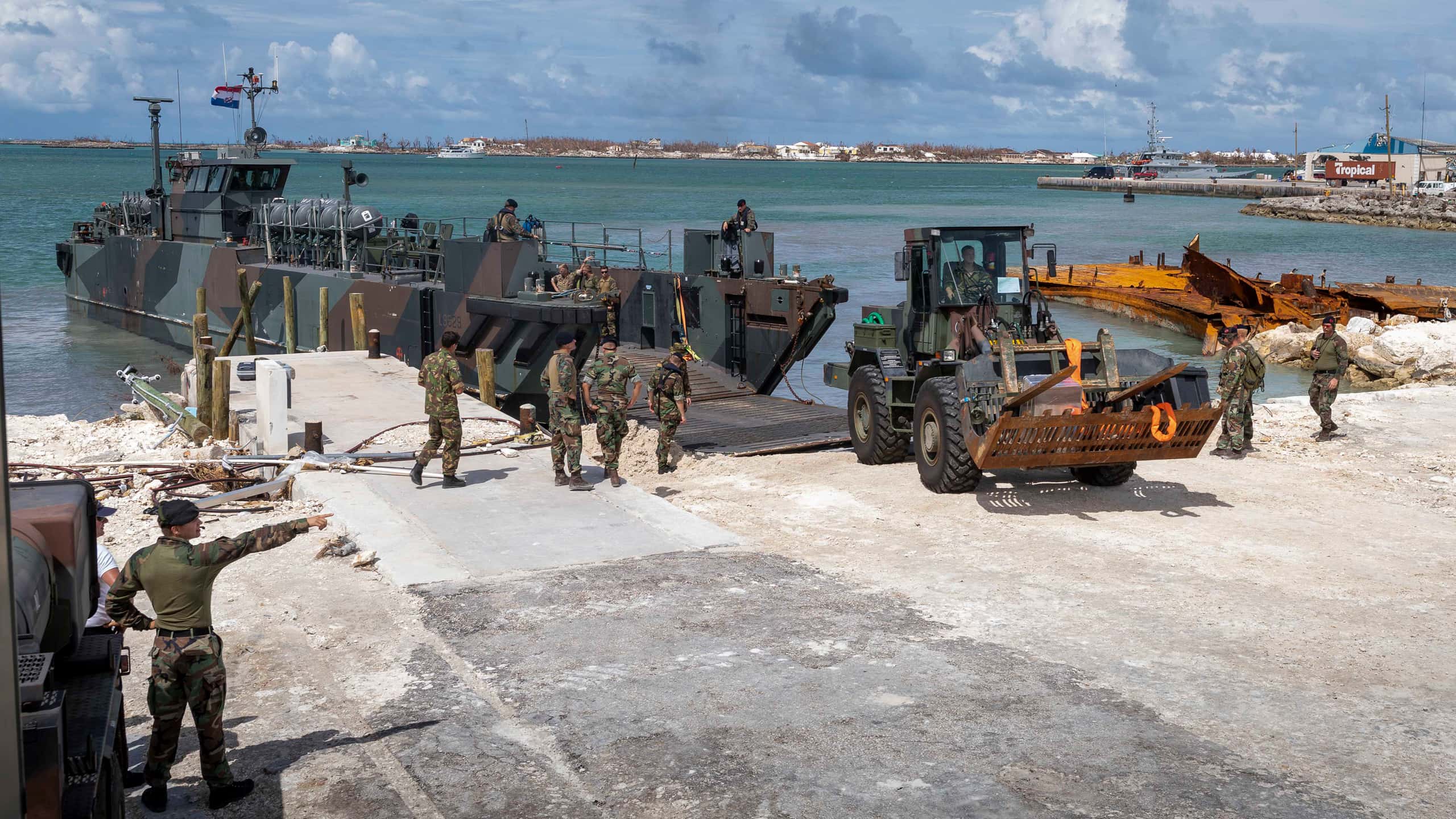 Kleurenfoto. Een militair landingsvaartuig is aan een haven aangemeerd en heeft de laadklep naar beneden. Een grote bulldozer rijdt de kant op. Op de kade staan meerdere militairen, gekleed in uniforms in camouflagekleding. Linksonder staat een militair die naar iets rechts in de verte wijst. In de achtergrond een havengebied.