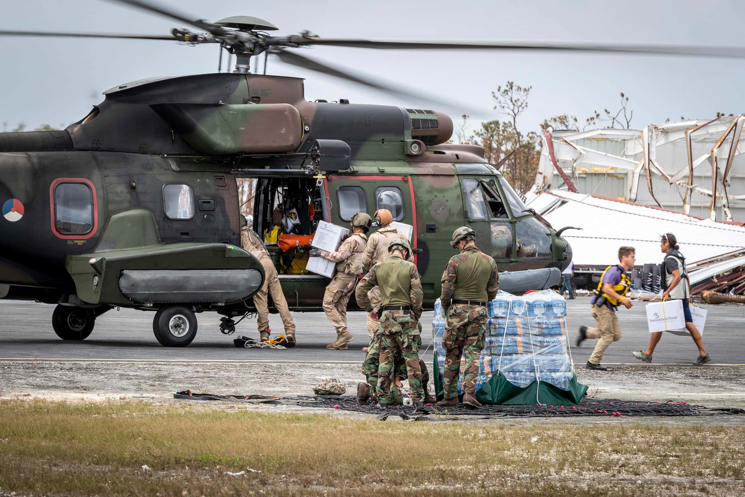Kleurenfoto. In de voorgrond staan drie militairen, gekleed in uniforms in camouflagekleuren, naast een stapel pakket drinkwater. Op de grond om de palet ligt een net. In de achtergrond staat een helikopter, in camouflagekleuren geschilderd, met openstaande zijdeur. Twee militairen laden dozen in, rechts rent iemand van de helikopter weg, terwijl een ander met twee dozen naar de helikopter loopt. In de achtergrond een ingestort gebouw.