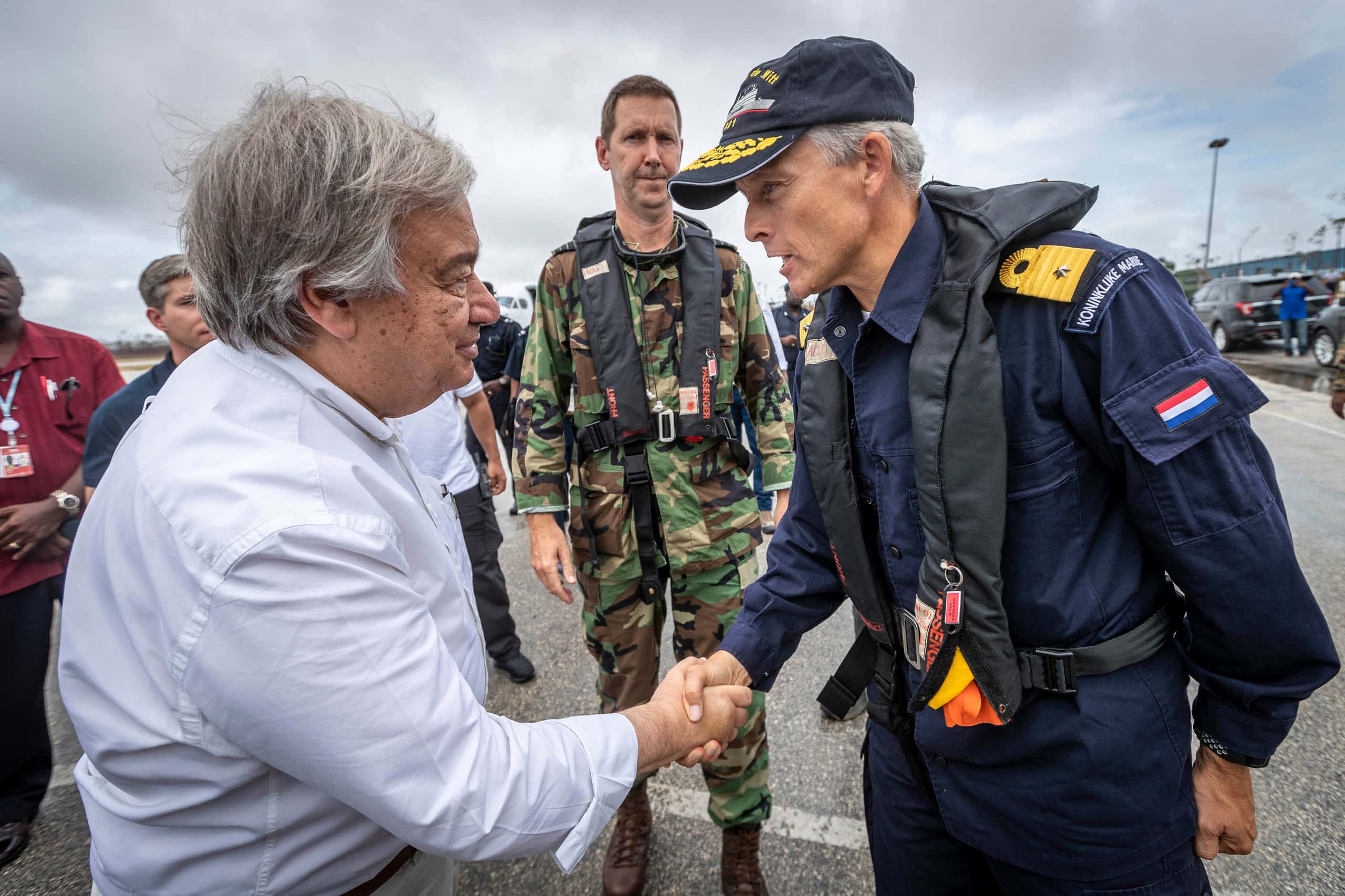 Kleurenfoto. Twee mannen staan tegenover elkaar en schudden elkaar de hand. De man rechts is militair en draagt een donkerblauw uniform, inclusief zwart reddingsvest en een pet. Op zijn schouder is een goudkleurig onderscheidingsteken te zien. De man links draagt een lichtblauw overhemd. In het midden staat een militair ineen uniform in camouflagekleuren, ook hij met een zwart reddingsvest.