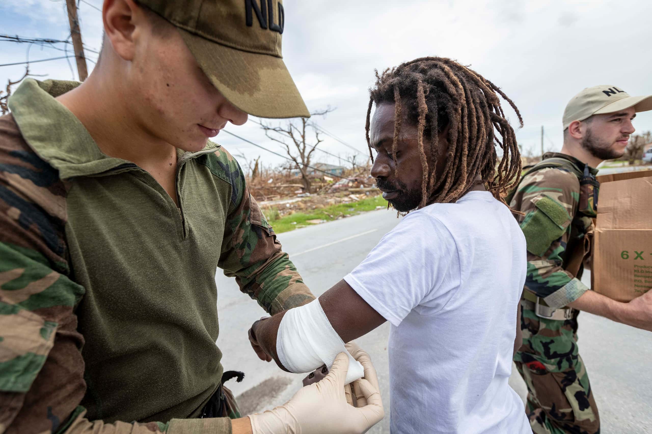 Kleurenfoto. Links staat een militair, gekleed in een uniform in camouflagekleding, met bezweette pet. Hij verbind de ellenboog van een man in een wit T-shirt en dreadlocks. Rechts draagt een andere militair een doos weg. In de achtergrond is veel puin te zien.