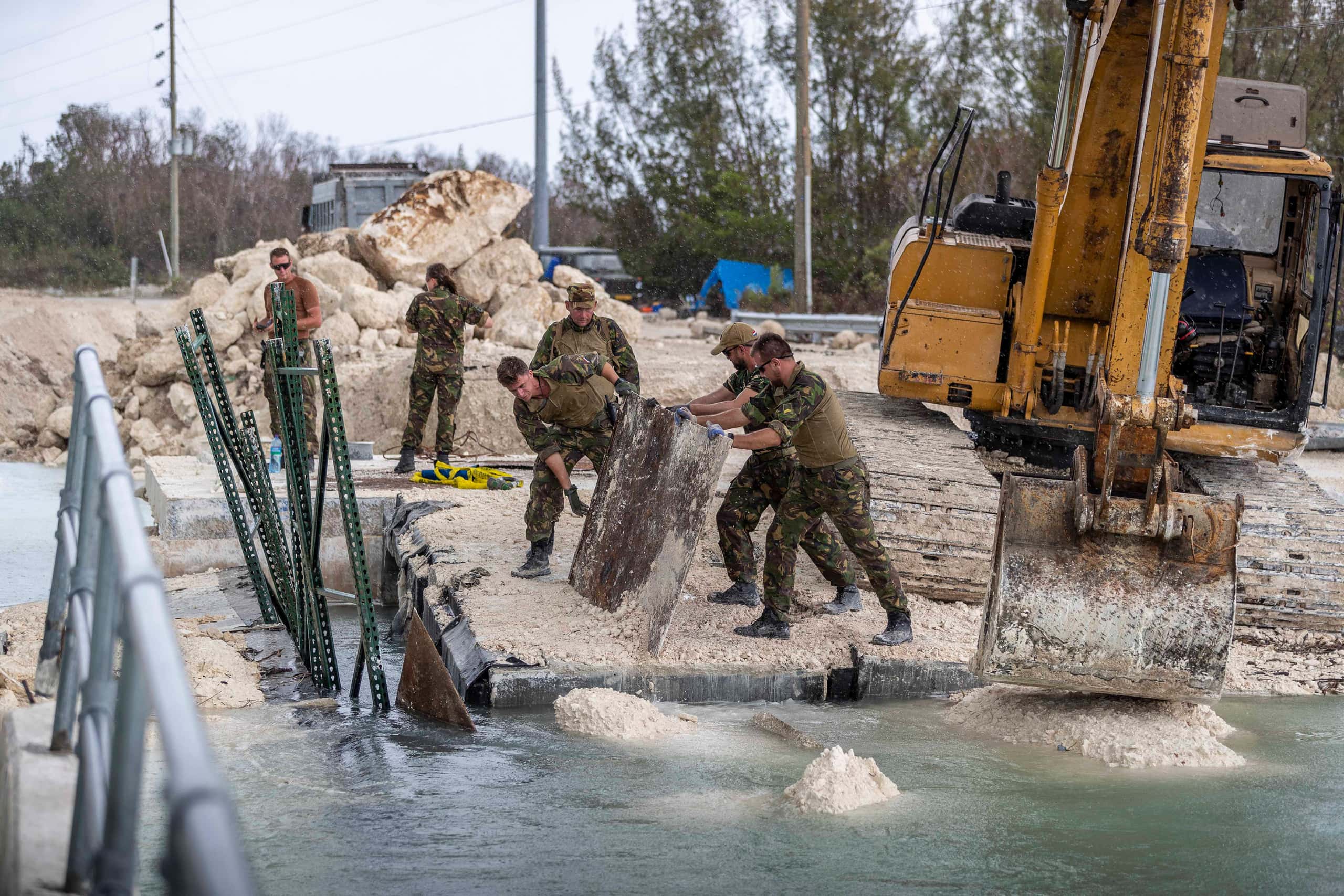 Kleurenfoto. Vier militairen staan op pontons in het water, de pontons liggen tegen de kant aan. Drie van hen schuiven met een grote metalen plaat zand en grind van de ponton. Rechts staat een grote graafmachine. Links van de ponton steken een aantal groene spijlen uit het water omhoog. Aan de kant staan nog twee militairen voor een stapel betonblokken.