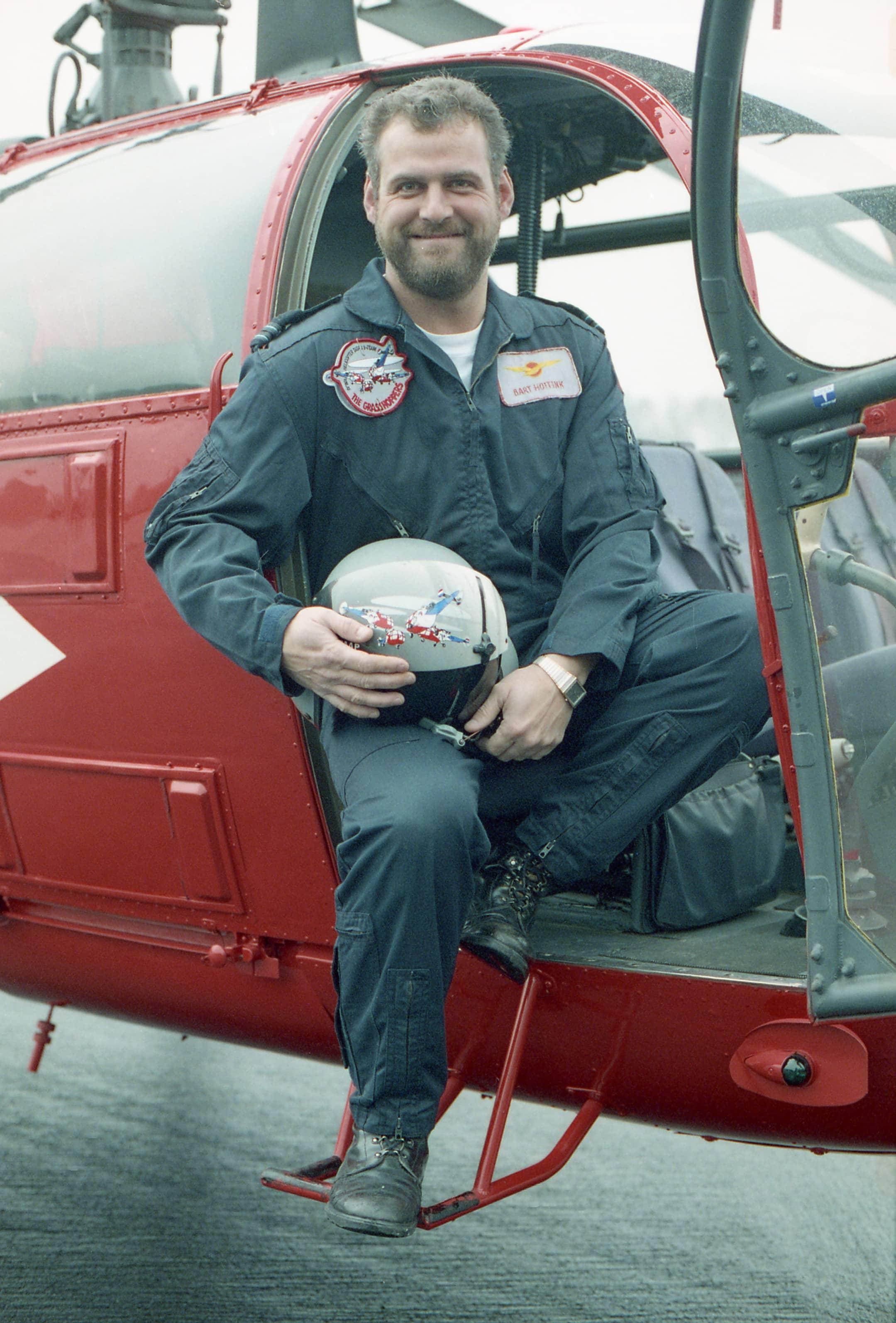 Kleurenfoto. Een man in een militaire overall poseert zittend in een rode helikopter. In zijn handen houdt hij een helm.