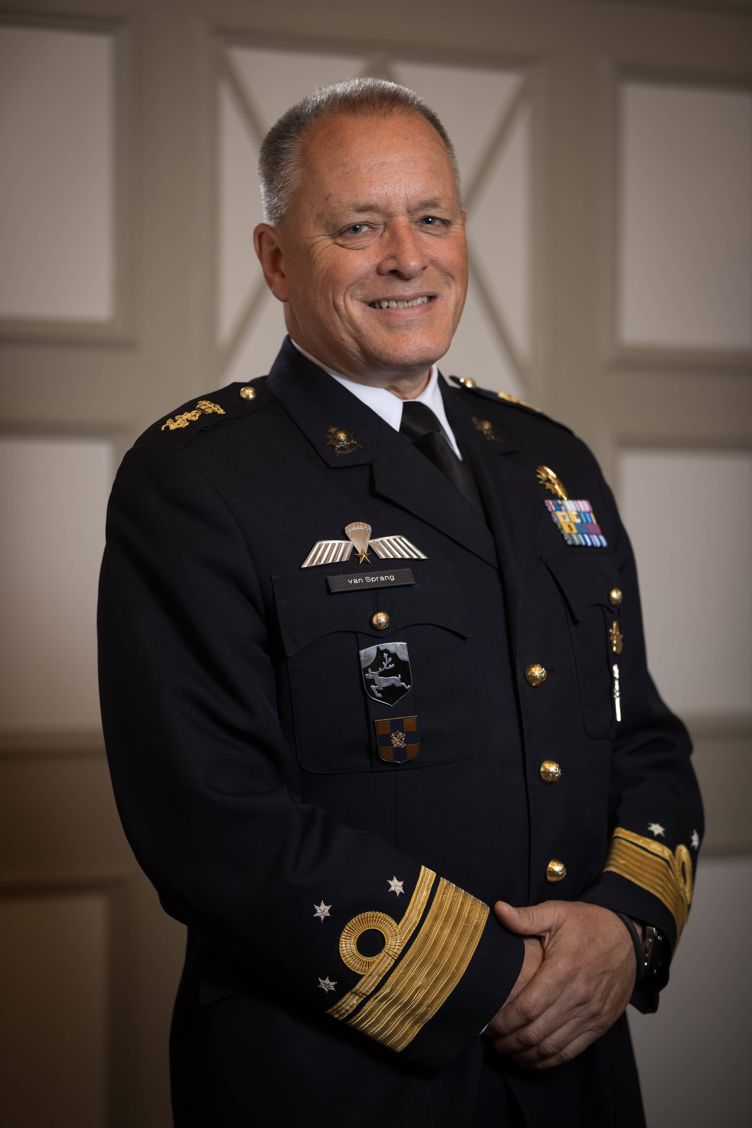 Kleurenfoto. Een man in donkerblauw uniform poseert voor de camera. Hij heeft zijn handen voor zich gevouwen. Hij staat voor een neutrale achtergrond.