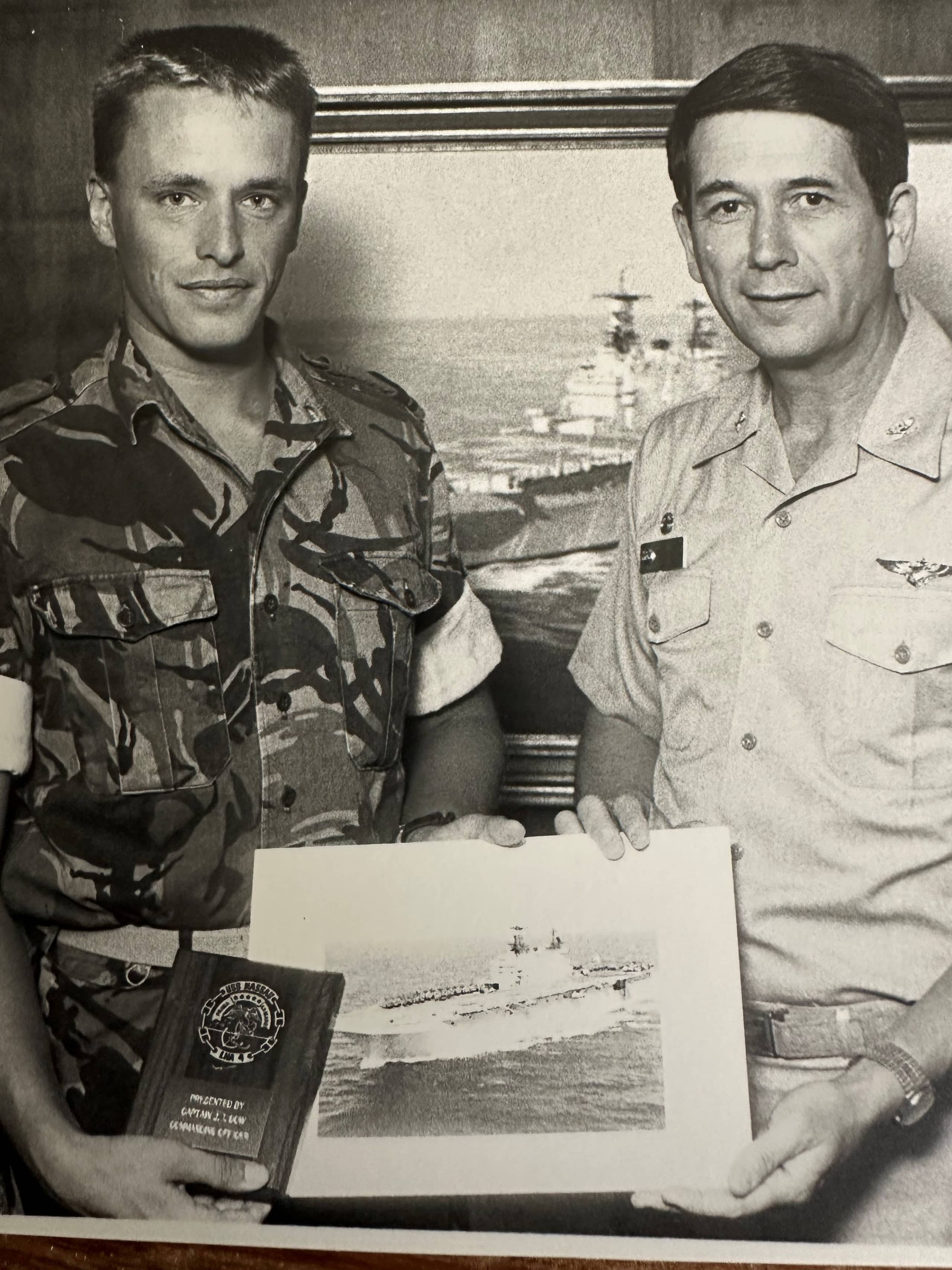 Zwart-wit foto. Twee mannen poseren staand voor de camera. Beiden zijn in militair uniform. Zij houden een foto van een marineschip vast. Op de achtergrond hangt een schilderij van een marineschip.