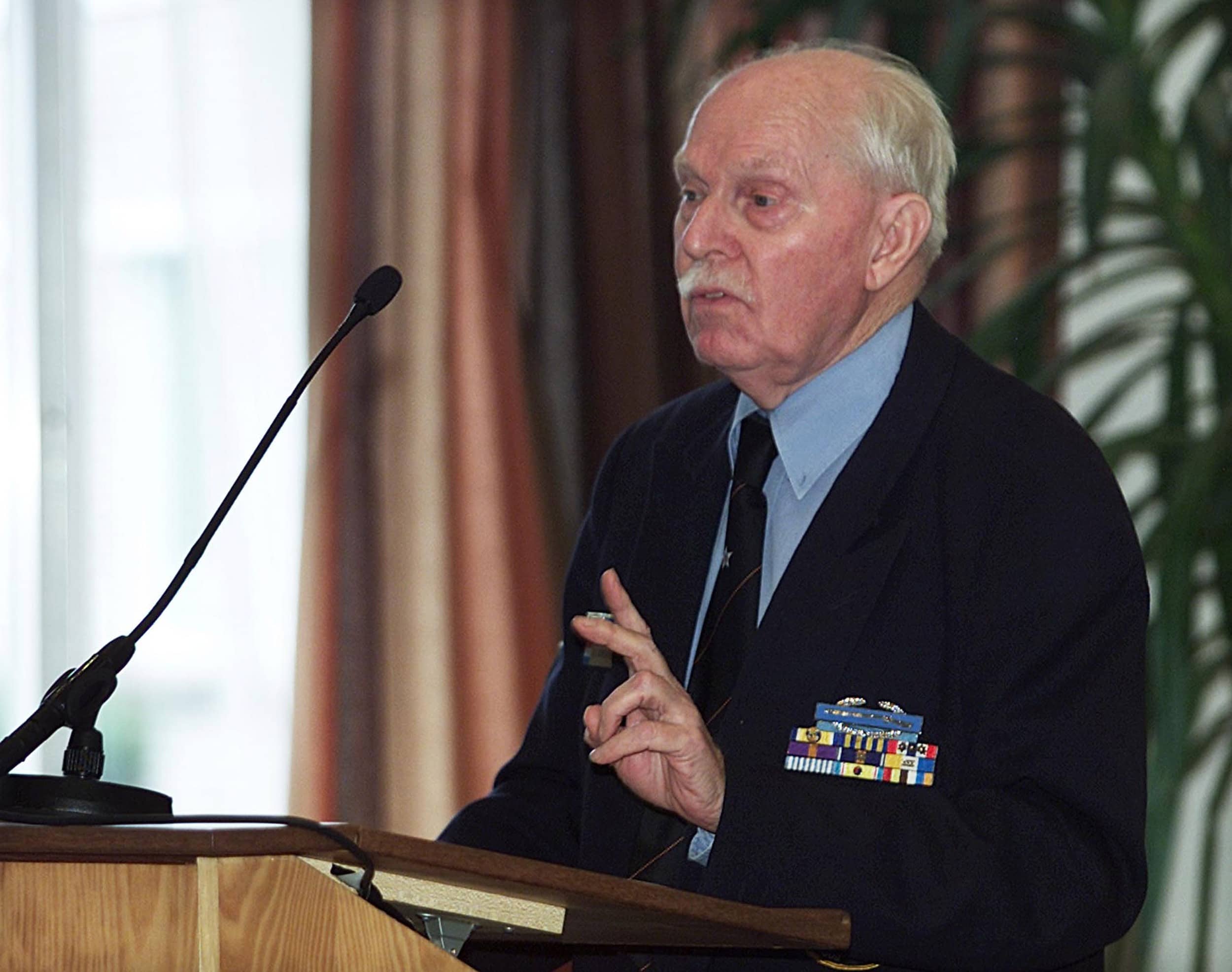 Kleurenfoto. Een man staat achter een lessenaar, hij praat en houdt twee vingers ophoog. Hij draagt een donker pak, lichtblauw overhemd en donkere stropdas, zijn haar en snor zijn grijs. Alleen de bovenste helft van zijn lichaam is te zien. Op zijn jasje prijken meerdere onderscheidingen. Links steekt vanuit de lessenaar een microfoon omhoog.
