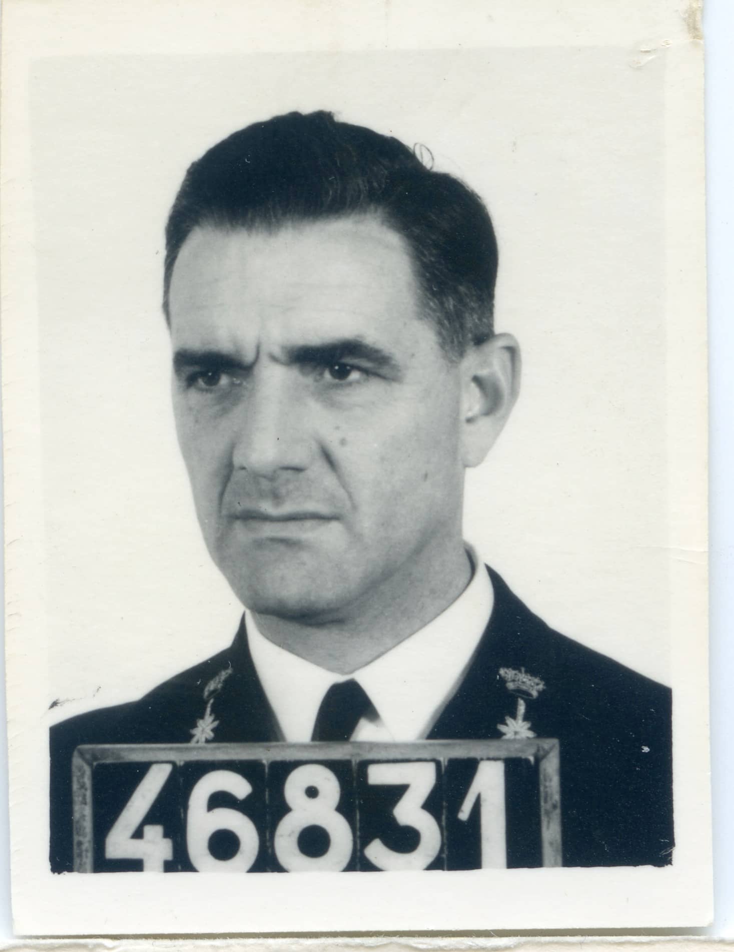 Zwart-witfoto. Dit is een portretfoto van een man. Het is nog net te zien dat hij een marine-uniformjas aanheeft, omdat op de boorden van zijn jas een kroon en daaronder het roer van een schip staat. Onder de jas zit een wit overhemd met een zwarte stropdas. Voor zich het bordje met zijn nummer, 46831. De man kijkt nors, schuin langs de camera. Hij heeft dik, donker haar dat schuin naar achteren is gekamd.