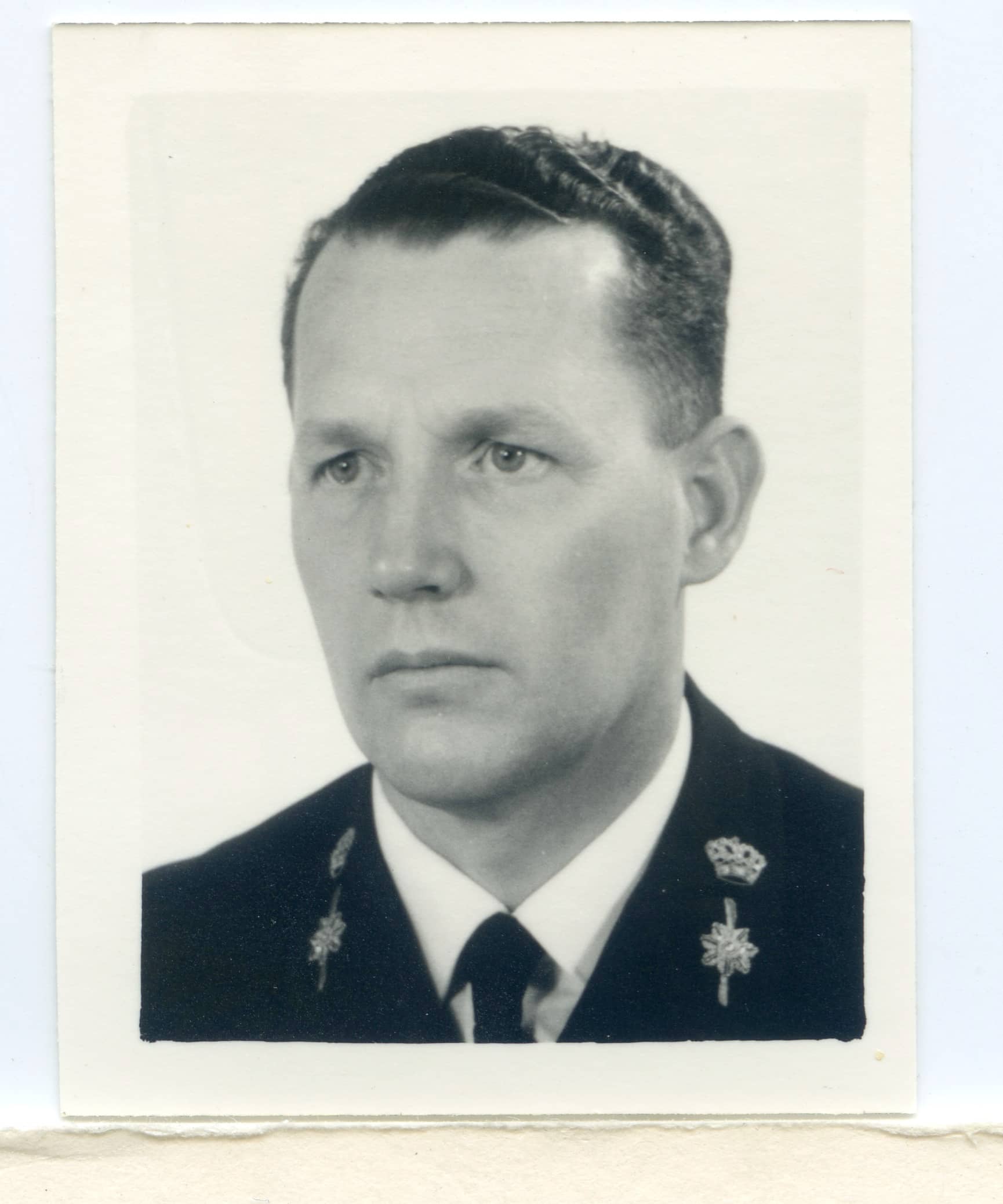 Zwart-witfoto. Dit is een portretfoto van een man. Het is nog net te zien dat hij een marine-uniformjas aanheeft, omdat op de boorden van zijn jas een kroon en daaronder het roer van een schip staat. Onder de jas zit een wit overhemd met een zwarte stropdas. De man kijkt serieus en zit schuin voor de camera. Hij heeft kort, donker, golvend haar.