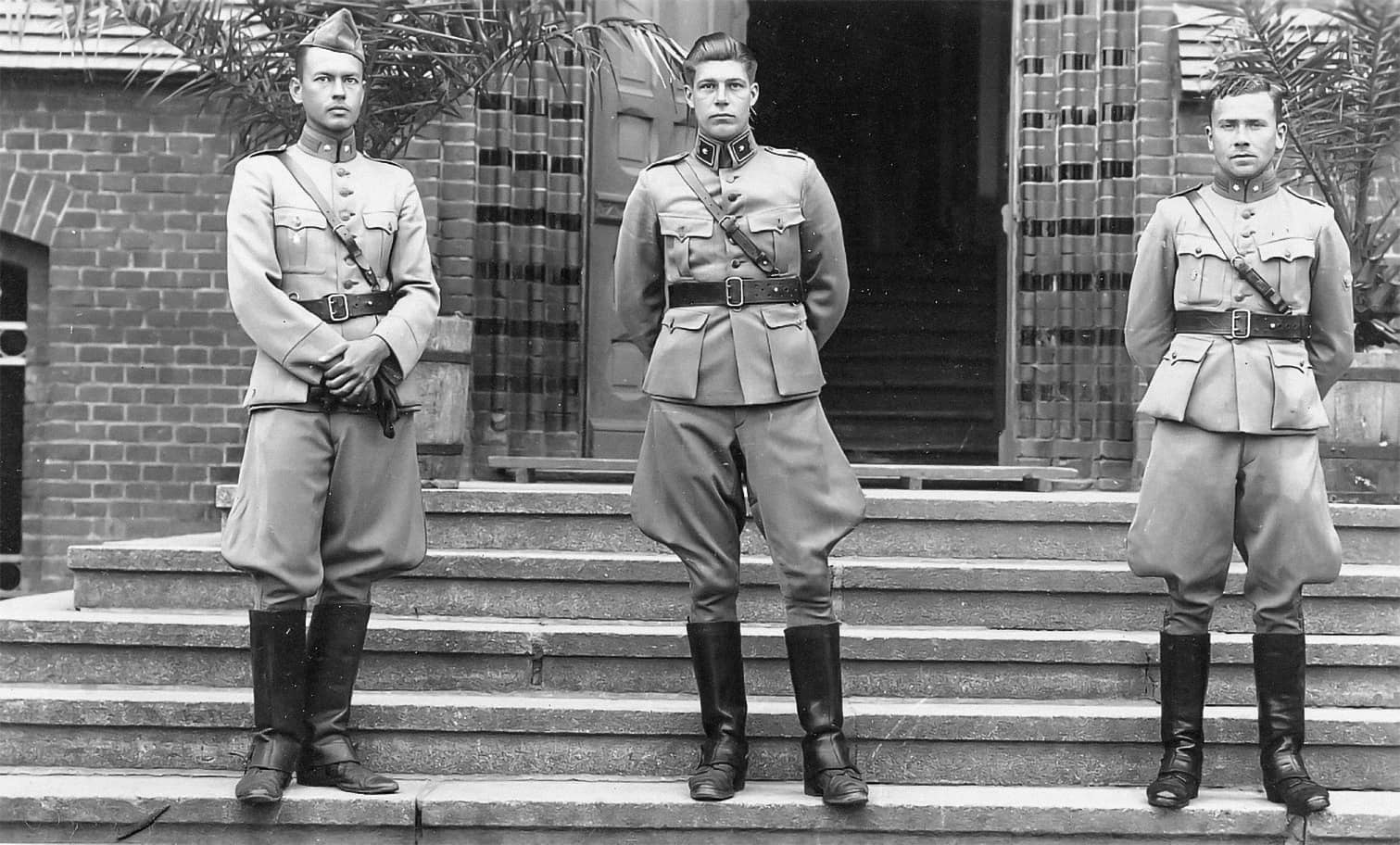 Zwart-witfoto. Drie mannen staan op het bordes voor een openstaande deur. Ze dragen alle drie militaire uniformen met wijde broeken, leren laarzen en riemen. Alle drie dragen ze een riem om hun middel en schuin over de borst.