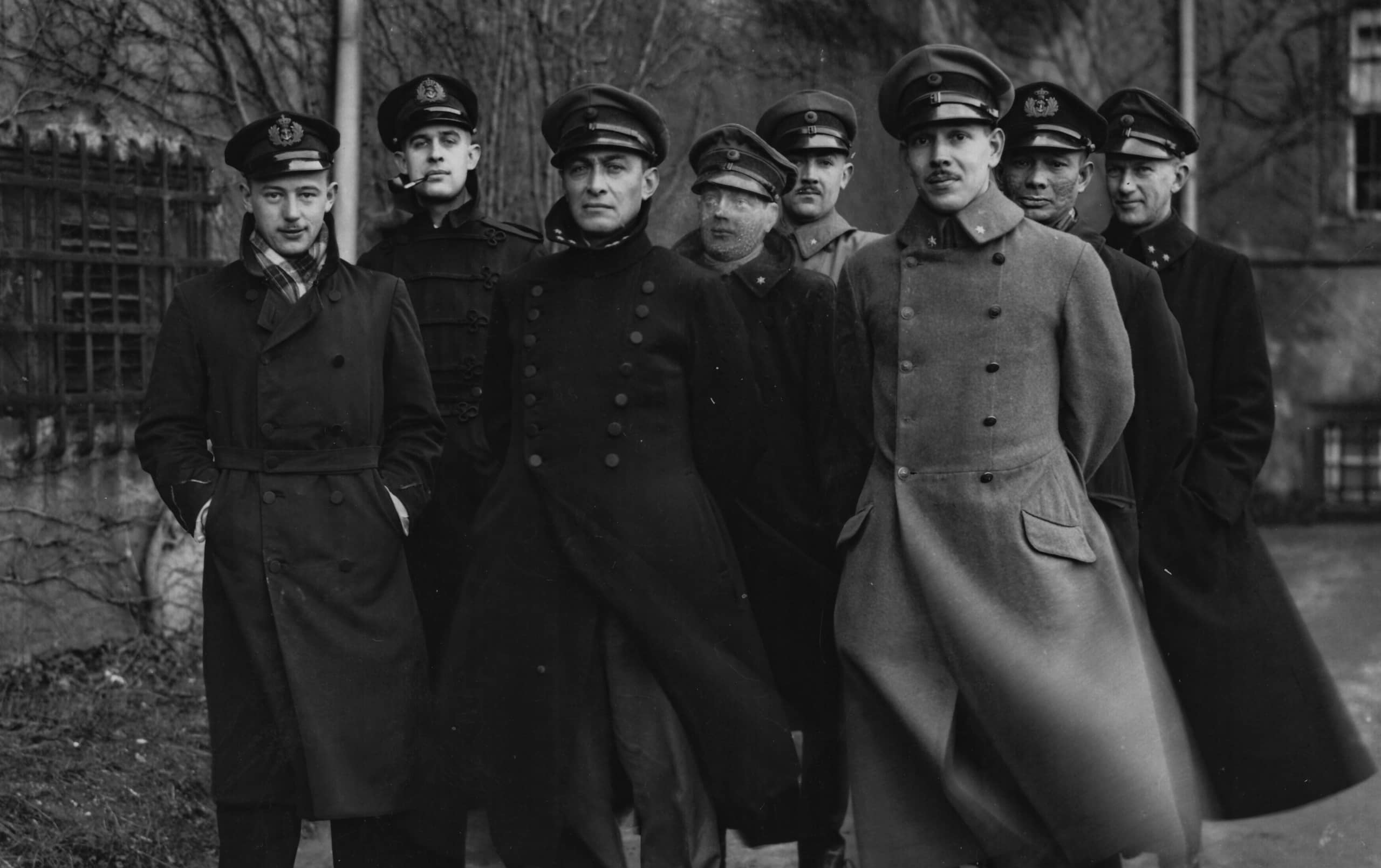 Zwart-witfoto. Een groep van zeven mannen staat in formatie, ze dragen allemaal lange overjassen. In het midden is het hoofd van een pop te zien, die verkleed is in een militair uniform.
