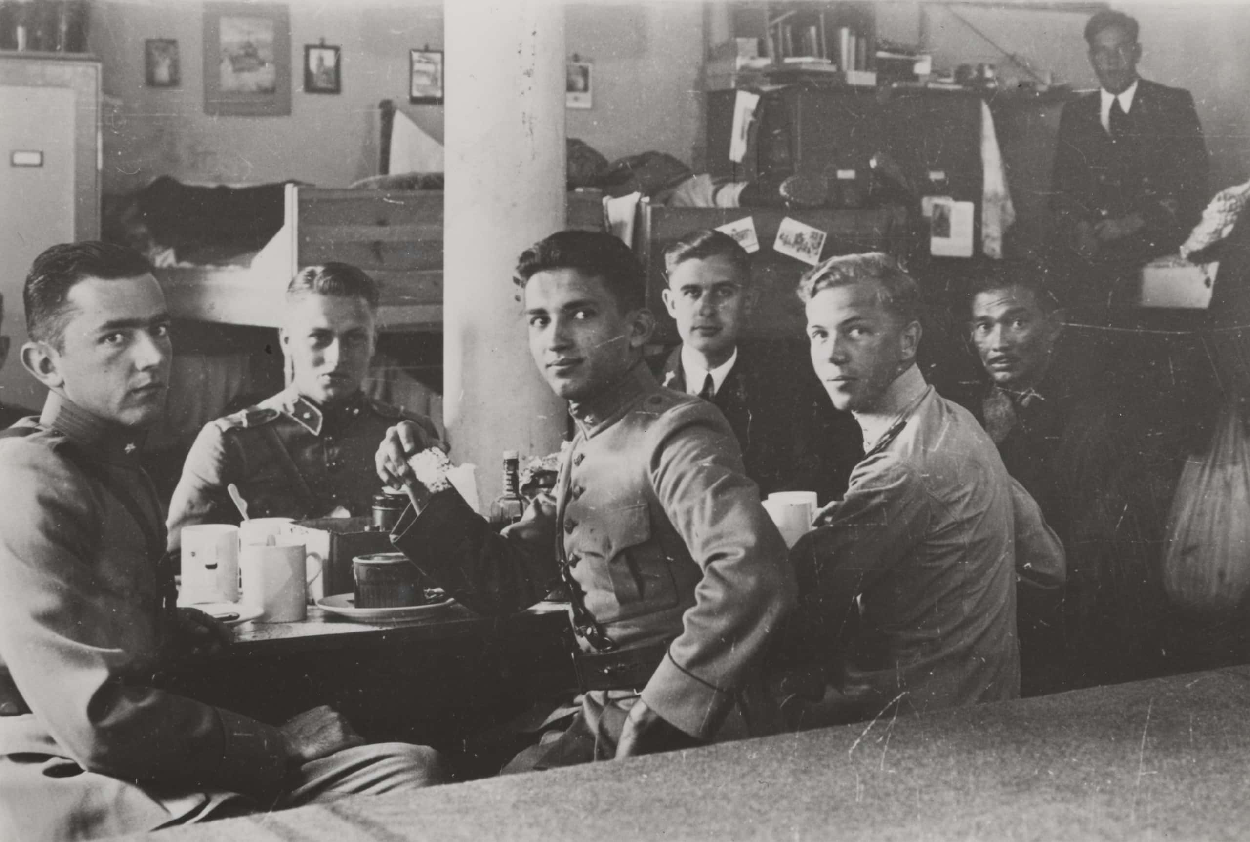 Zwart-witfoto. Foto van zes militairen die aan een tafel zitten. Drie kijken half gedraaid achterom naar de camera. Ze dragen allemaal militaire uniforms. Op tafel staan flessen, borden en mokken. In de achtergrond een paar stapelbedden en kasten met boeken erop. Rechts in de achtergrond staat een zevende militair. Aan de muur hangen fotolijstjes.