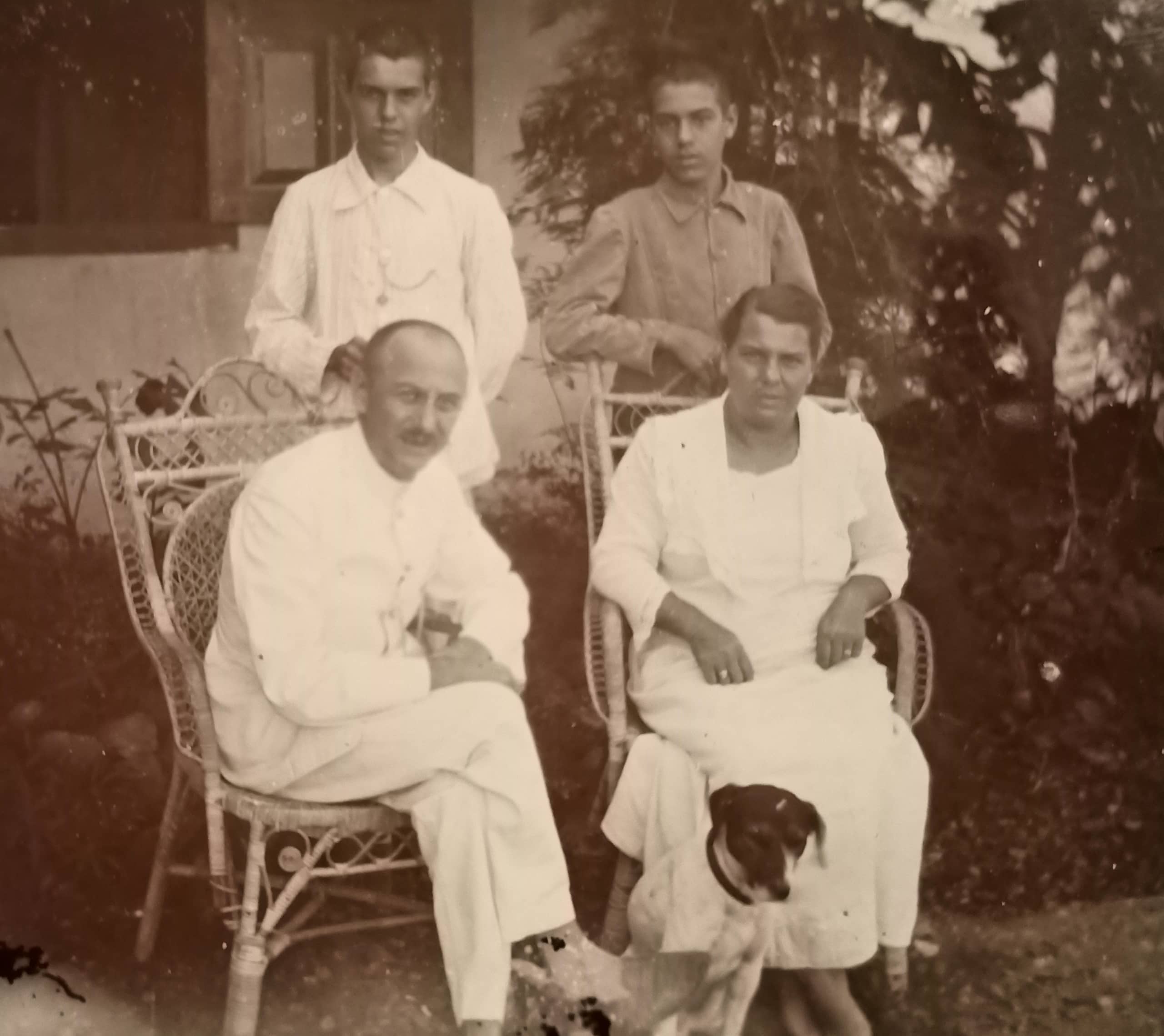 Zwart-witfoto. Portret van een gezin. De man en vrouw zitten op rieten stoelen, daarachter staan twee jongens. De man, vrouw en de linker jongen dragen witte kleding, de rechter jongen draagt een donkerder hemd. Voor de vrouw zit een hond. In de achtergrond struiken van een tuin en de gevel van een huis.