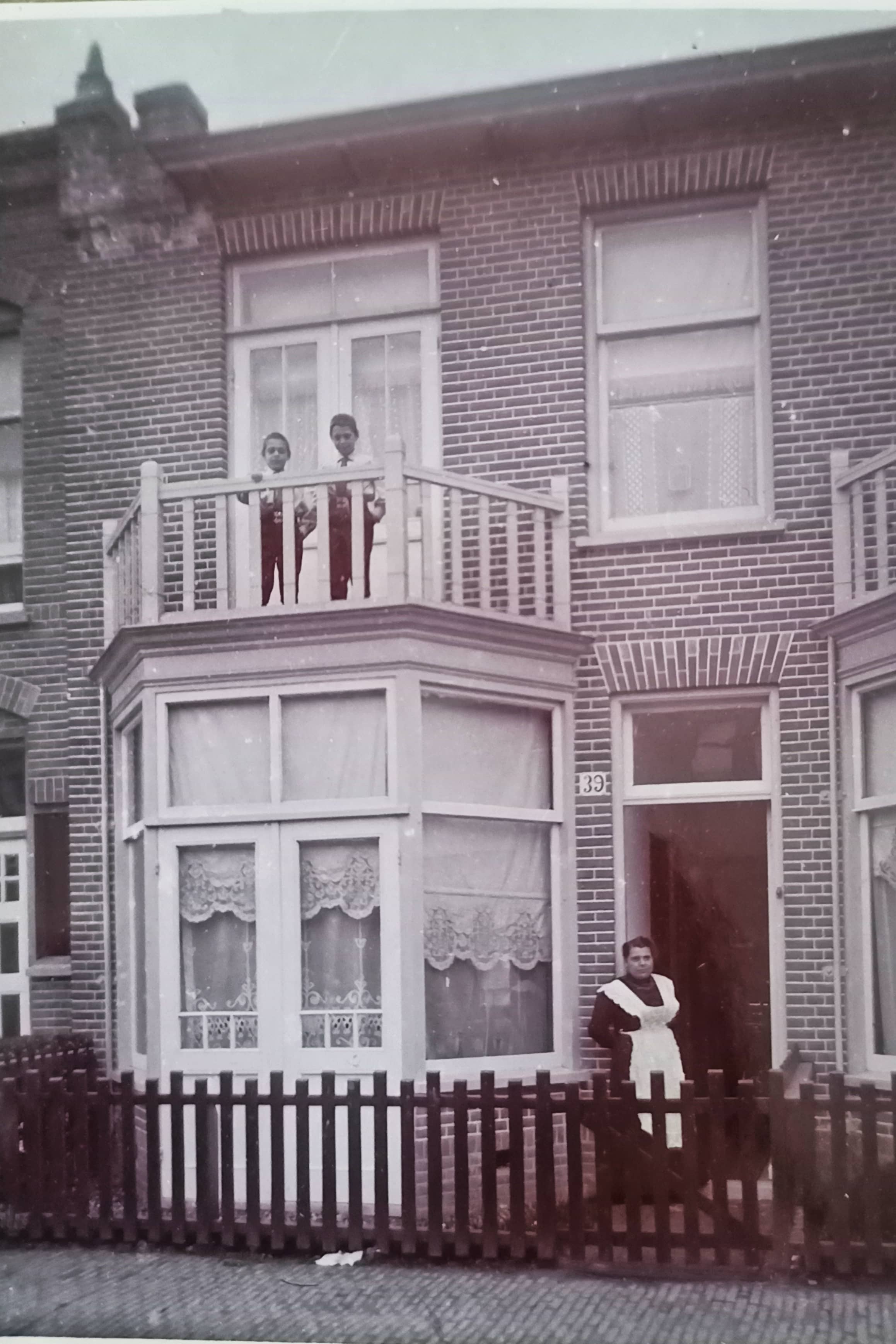 Zwart-witfoto. Foto van een rijtjeshuis met een kleine voortuin. Bij de openstaande voordeur staat een dienstmeisje, zij draagt een zwarte jurk met witte schort. Het huisnummer is 39. Voor de ramen van de uitbouw op de begane grond hangen witte gordijnen Op het balkon staan twee jongetjes.