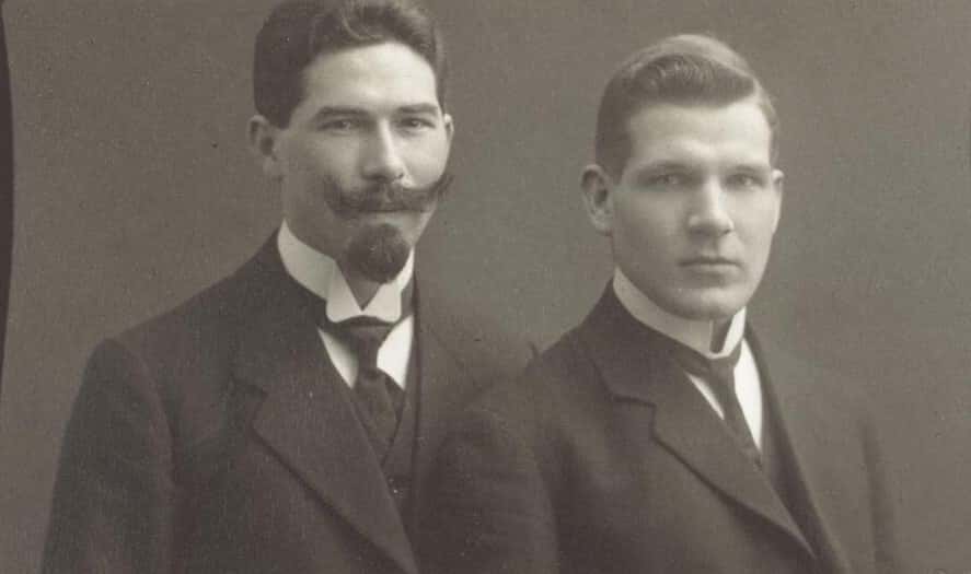 Zwart-witfoto. Twee mannen samen op de foto. De man links draagt een donker pak met donkere das en gilet. Zijn haar is zwart, hij heeft een snor met borstelige, opstaande uiteinden en een sik. De man rechts draagt dezelfde kleding en heeft geen gezichtsbeharing.