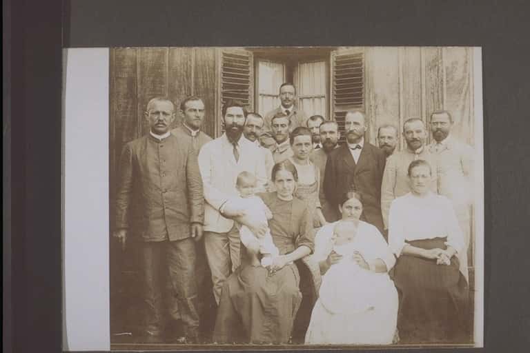 Zwart-witfoto. Groepsfoto van achttien personen: vier vrouwen, twee baby’s en verder mannen. Drie vrouwen zitten op de eerste rij, twee hebben de baby’s vast, de rest staat erachter. Links een man in een wit pak en zwarte baard. In de achtergrond een houten huis met raam.