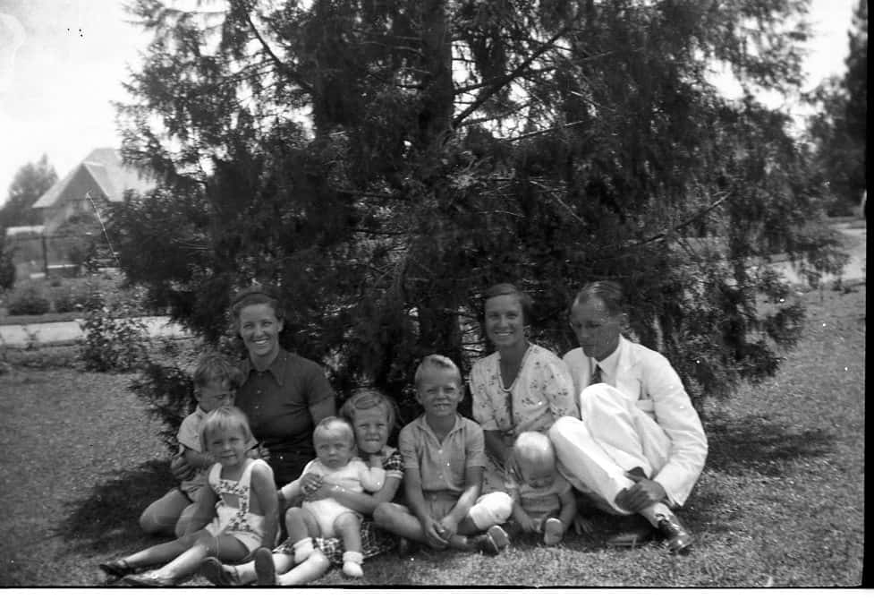 Zwart-witfoto. Drie volwassenen, twee vrouwen en een man, zitten in de schaduw van een boom. Voor en tussen hen in zitten zes kinderen: twee jongens en vier meisjes. De man zit helemaal rechts, draagt een wit pak en houdt met zijn hand beide enkels vast. Hij kijkt niet in de camera maar bezorgd of afgeleid naar de grond.