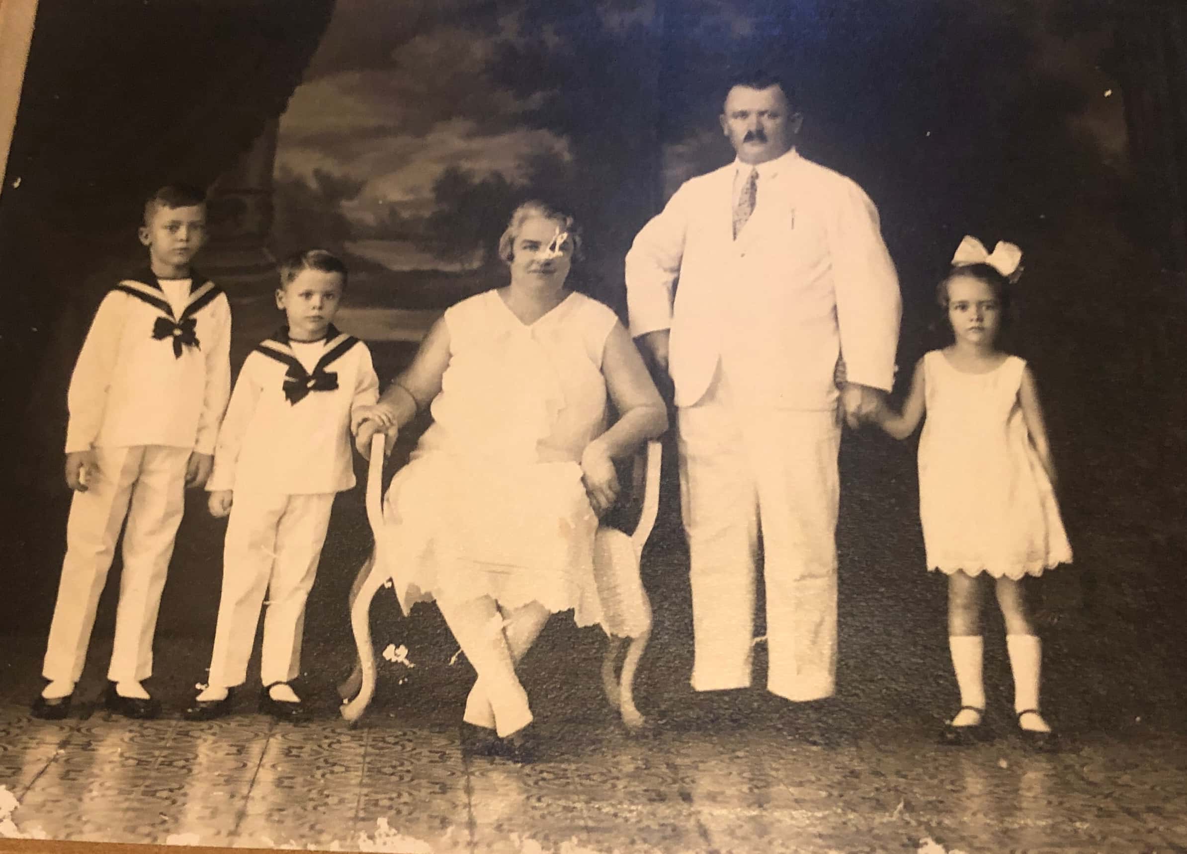 Zwart-witfoto. Een gezin van drie kinderen en twee volwassenen poseert voor een foto. Links twee jongens, een vrouw zit prominent in een stoel in het midden, rechts van haar de man die de hand van een meisje vasthoudt. De jongens dragen matrozenpakjes, de anderen dragen witte kleding.