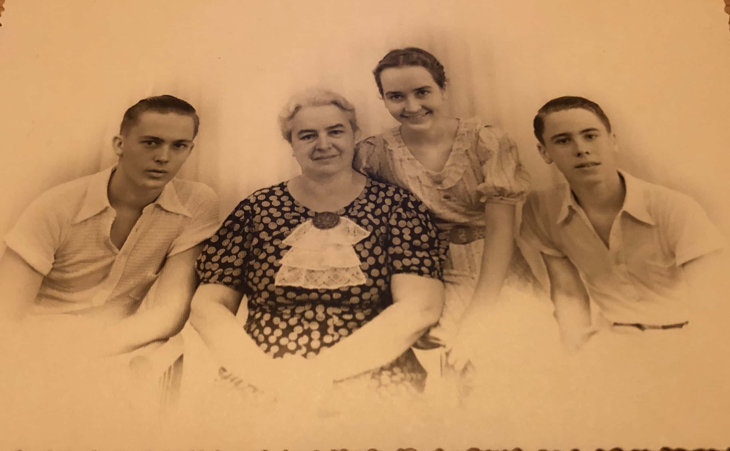 Zwart-witfoto. Een vrouw, een jongedame en twee jongemannen poseren voor een foto. De vrouw zit in het midden en draagt een donkere jurk met stippen, de jongedame zit rechts half op een armleuning. Naast hen zitten de twee jonge mannen. De jongeren dragen zomerse kleding.