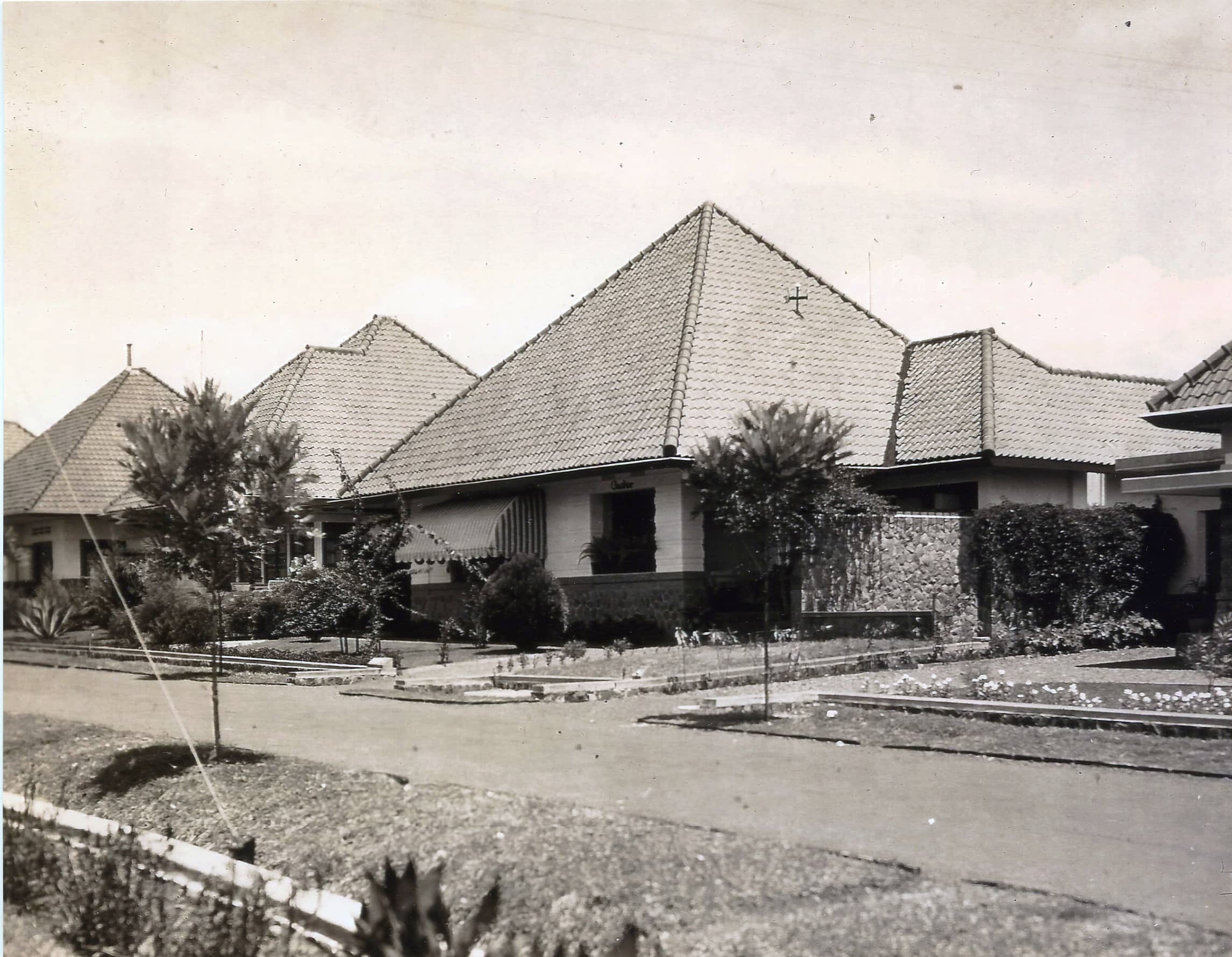 Zwart-witfoto. Straatbeeld met een rij vrijstaande huizen. Langs de weg staan jonge boompjes, in de voortuinen groeien bloemen en ligt gras. De huizen hebben lage muren en hoge piramidedaken. Het huis in het midden heeft de markies voor de ramen naar beneden hangen.