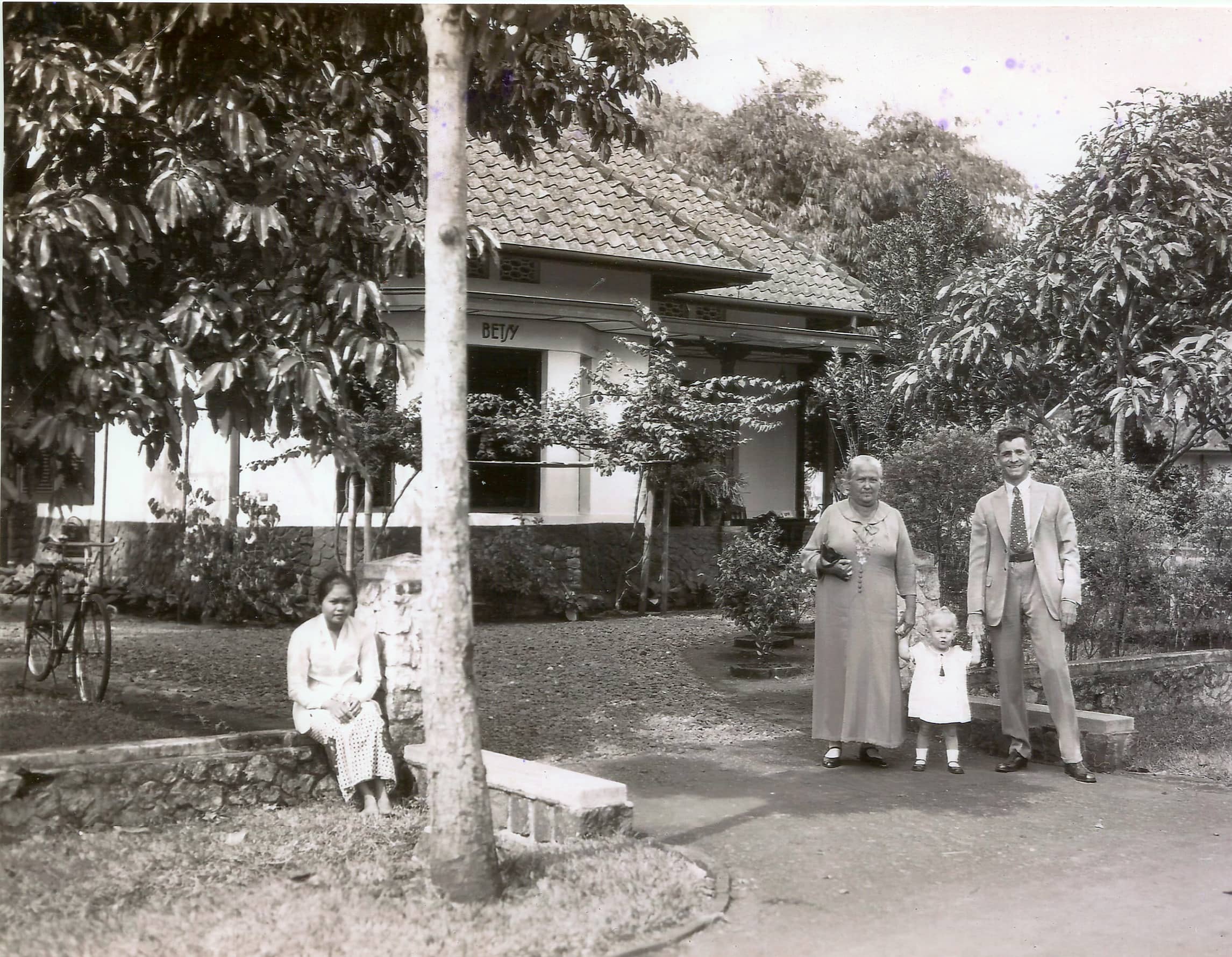 Zwart-witfoto. Rechts staan een man, klein meisje en oudere vrouw op een oprit naar een huis achter hen. De man en de vrouw houden de handen van het meisje vast dat tussen hen in staat. Het meisje draagt een wit jurkje. Links van de oprit zit een andere vrouw op een laag muurtje. De muren van het huis zijn wit en op de voorgevel is de naam Betsy geschilderd.