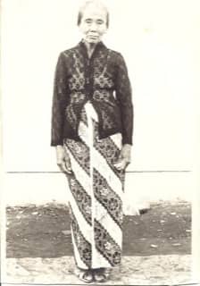 Zwart-witfoto. Foto van een vrouw, ze staat ergens in een straat en draagt traditionele Indonesische kleding.
