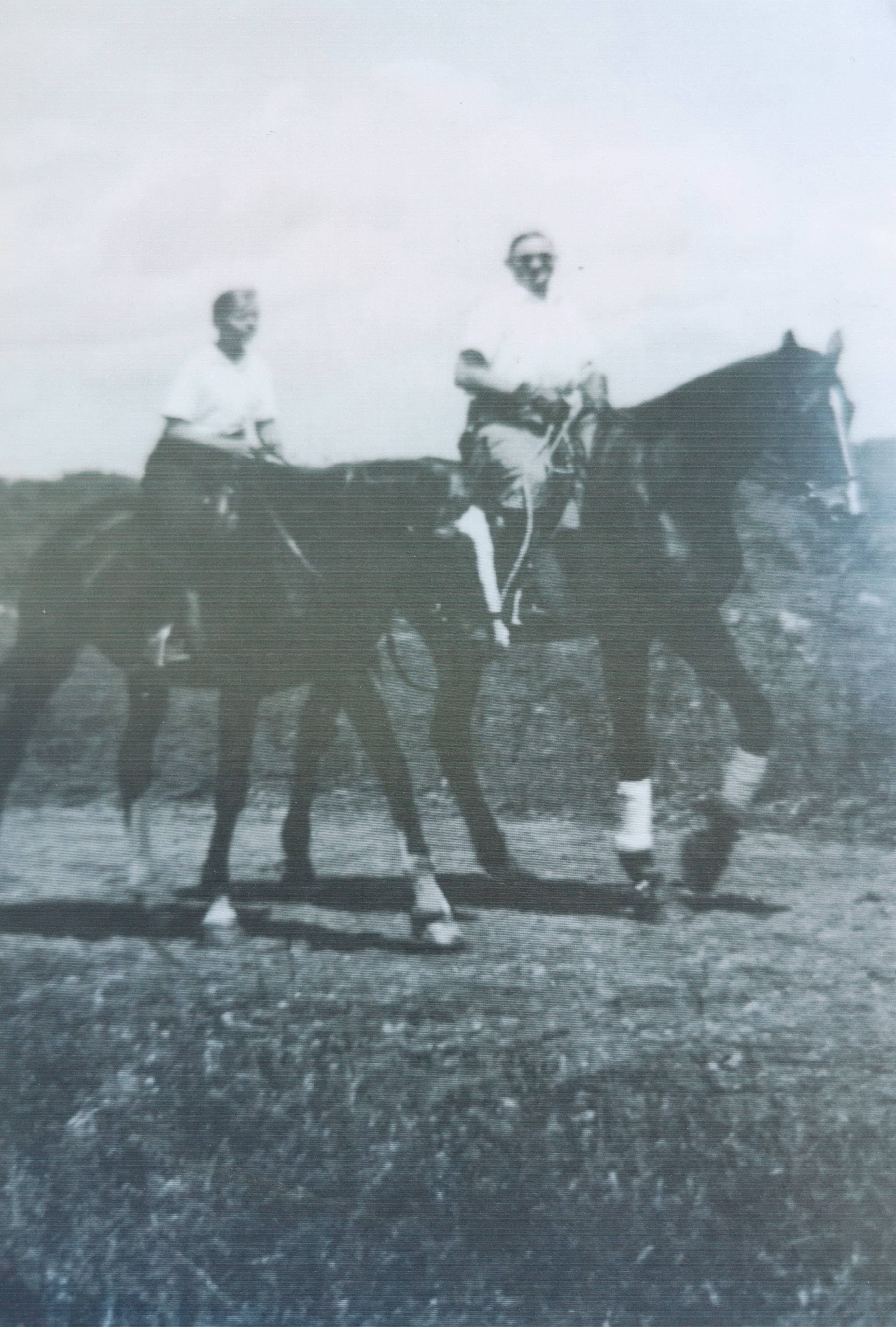 Zwart-witfoto. Te zien zijn twee paarden met daarop een jongeman (links) en een oudere man. De paarden zijn donker van kleur, met lichte sokken. Beide mannen dragen een licht hemd en donkere broek, de man rechts draagt ook een zonnebril en lacht naar de camera.