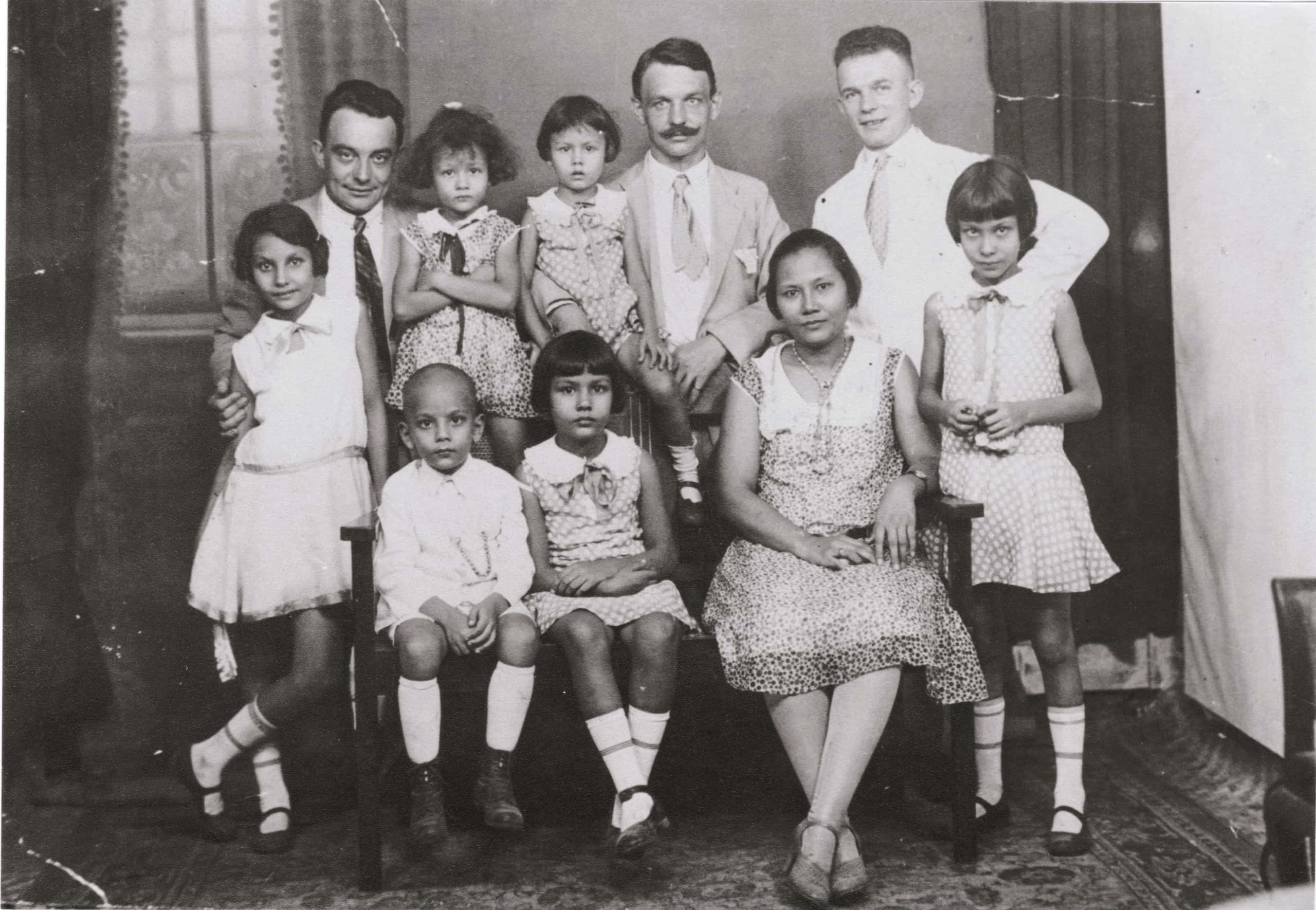 Zwart-witfoto. Groepsfoto van een gezin met zes kinderen: vijf meisjes en een jongen. De jongen zich links vooraan op een bankje, daarnaast een meisje en een vrouw. Achter het bankje staan drie mannen, de middelste heeft een snor met opstaande punten, zij dragen alle drie een pak met stropdas. Ook de kinderen en de vrouw zijn netjes gekleed.