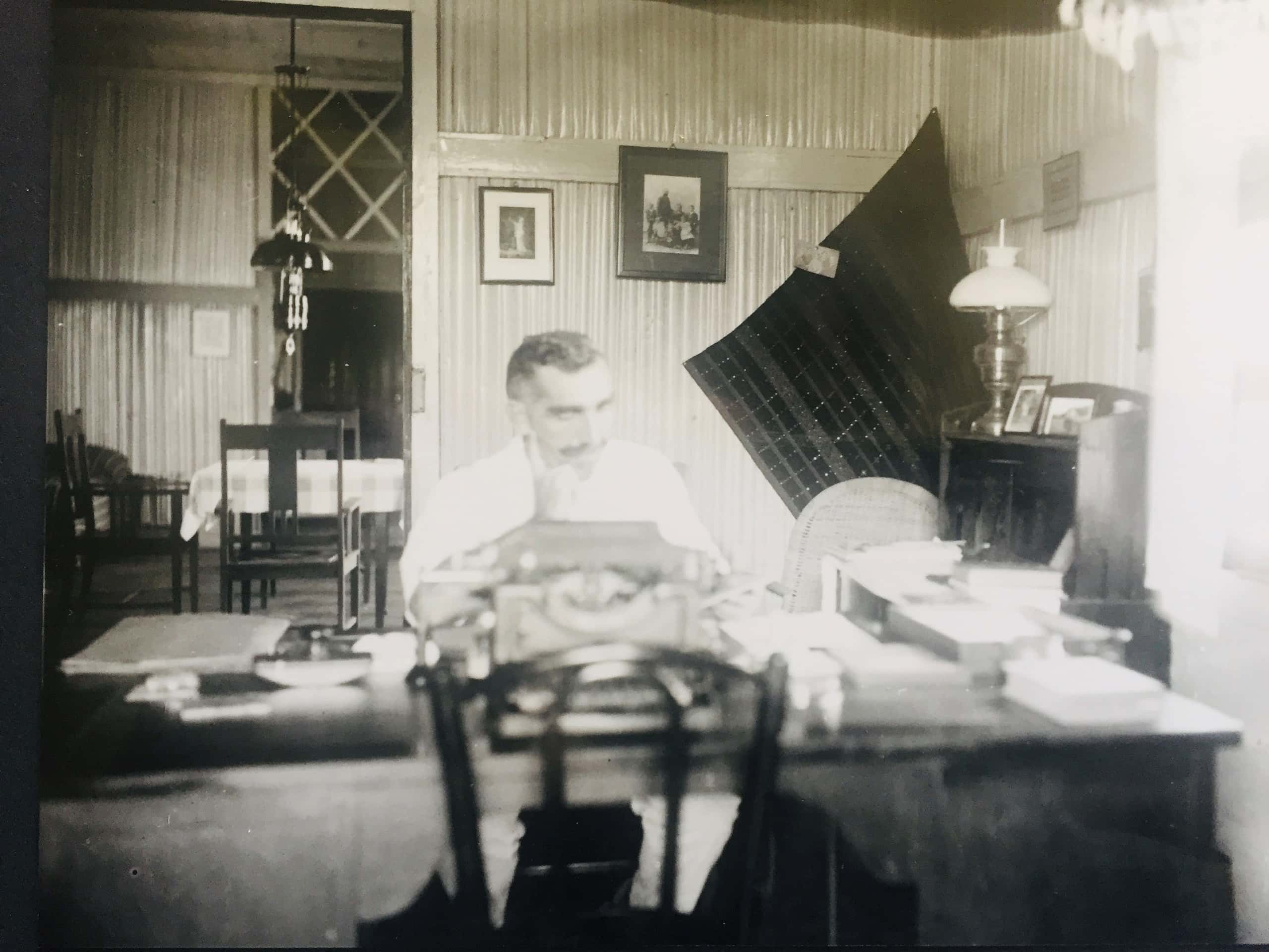 Zwart-witfoto. Een man zich achter een bureau, hij leunt met zijn kin op zijn rechterhand. Op het bureau liggen kantoorattributen, zoals een typemachine, mappen en papieren. Aan de muur achter hem hangen een paar fotolijsten en een wandtapijt, rechts tegen de muur staat een kastje. Links in de achtergrond een andere kamer met tafel en stoelen.