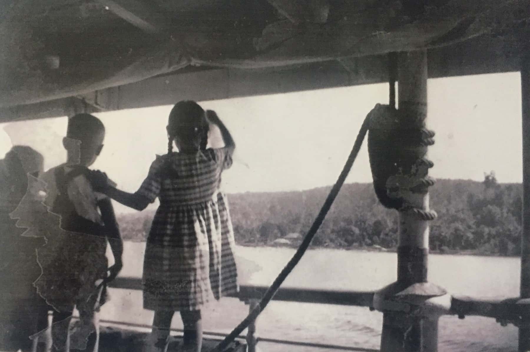 Zwart-witfoto. Drie kinderen, twee jongens en een meisje, staan op het dek van een schip en kijken naar de kust in de verte. Ze staan met de rug naar de camera, het meisje heeft haar linkerhand op de schouder van de jongen naast haar. Rechts een paal die het dak ondersteunt, er is een touw omheen geknoopt.