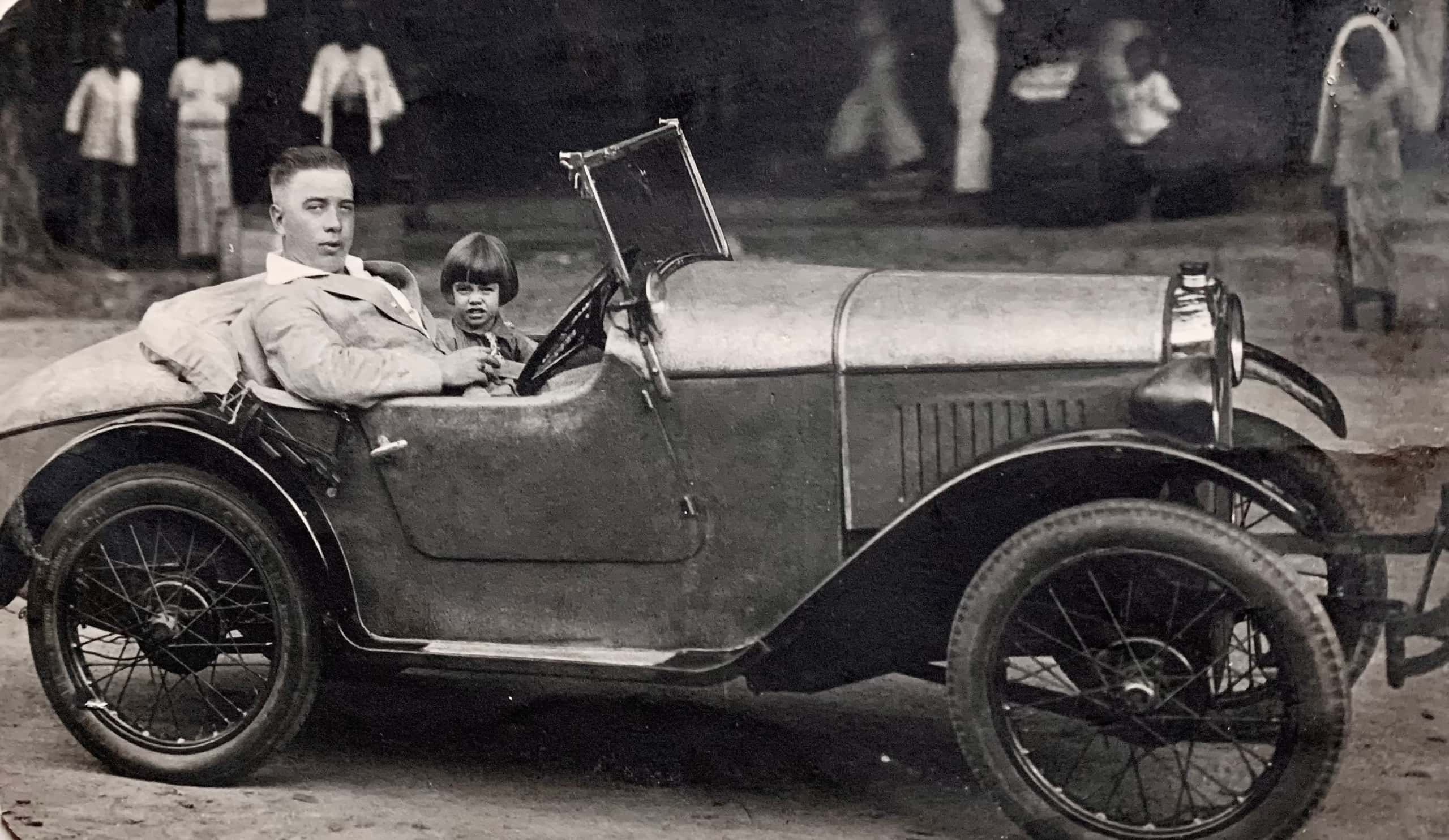 Zwart-witfoto. Een man zit ietwat achterover achter het stuur van een klassieke auto zonder dak. Hij draagt een licht pak en heeft een sigaret tussen zijn vingers. Naast hem zit een jong meisje. In de achtergrond een straatbeeld met mensen die naar de auto staan te kijken.