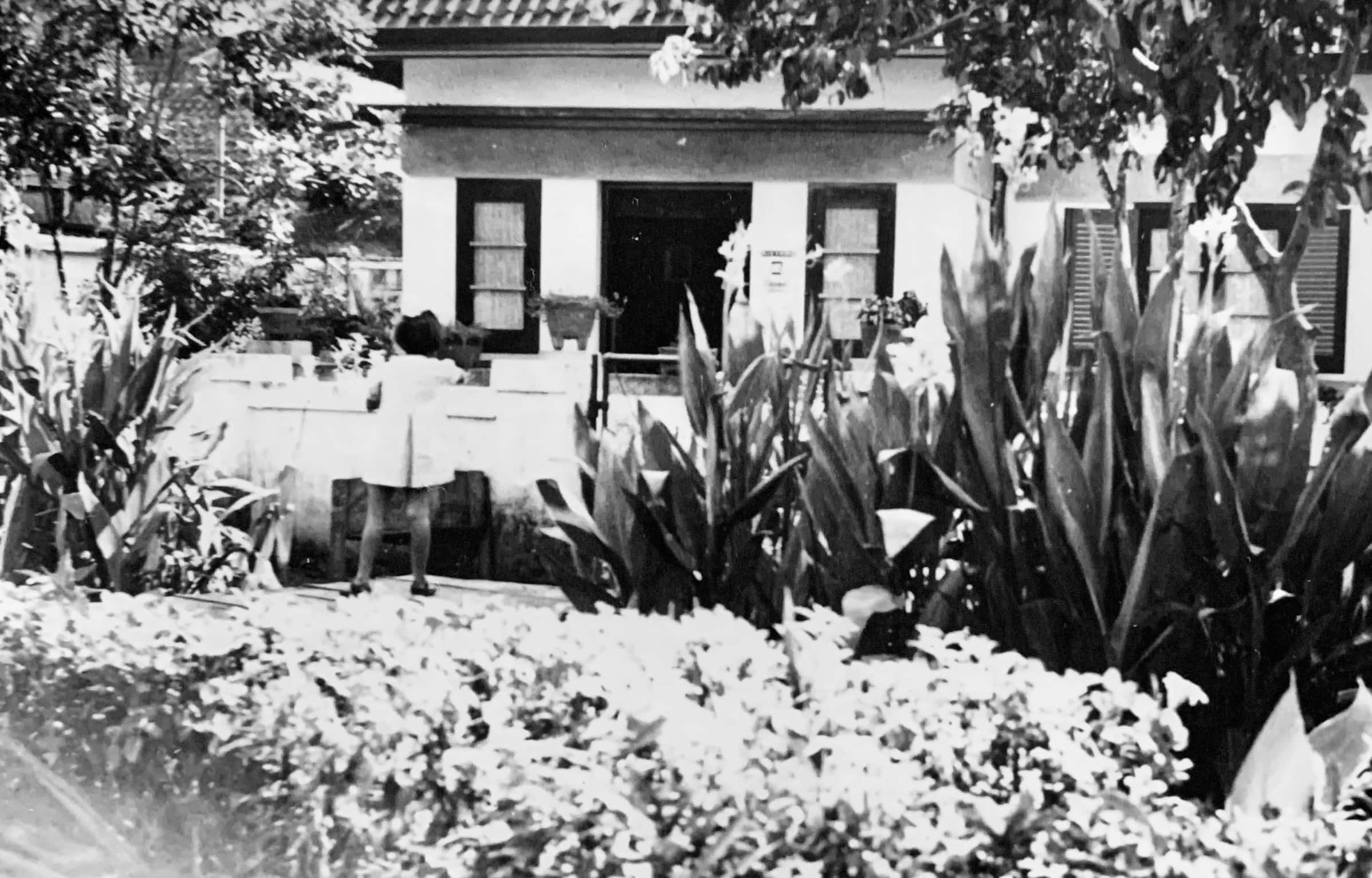 Zwart-witfoto. Vage foto van een tuin en een huis. Op de voorgrond lage struiken, daarachter hoge planten. Links van het midden staat een meisje in een witte jurk, zij staat met de rug naar de camera. In de achtergrond het huis en enkele bomen.