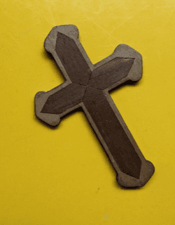 Kleurenfoto. Een bruin kruis van hout op een gele achtergrond. Het zijn twee kruisen in één. Het kruis aan de buitenkant heeft een lichtere kleur en heeft afgeronde uiteinden. Het binnenste kruis is donkerder van kleur, de vier uiteinden lopen in een punt.