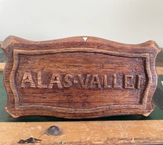 Kleurenfoto. Foto van een houten deksel of houten plaat. In het midden staat Alas-vallei geschreven, de tekst is omrand. Aan de korte kanten zit er een kleine knik in de omranding.