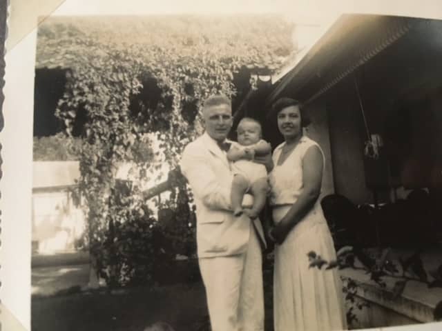 Zwart-witfoto. Een man en een vrouw staan in een tuin en poseren voor de foto, de man heeft een slapende baby in zijn armen. De man draagt een wit pak met donkere strik, de vrouw een witte jurk. In de achtergrond bomen van de tuin en een huis.